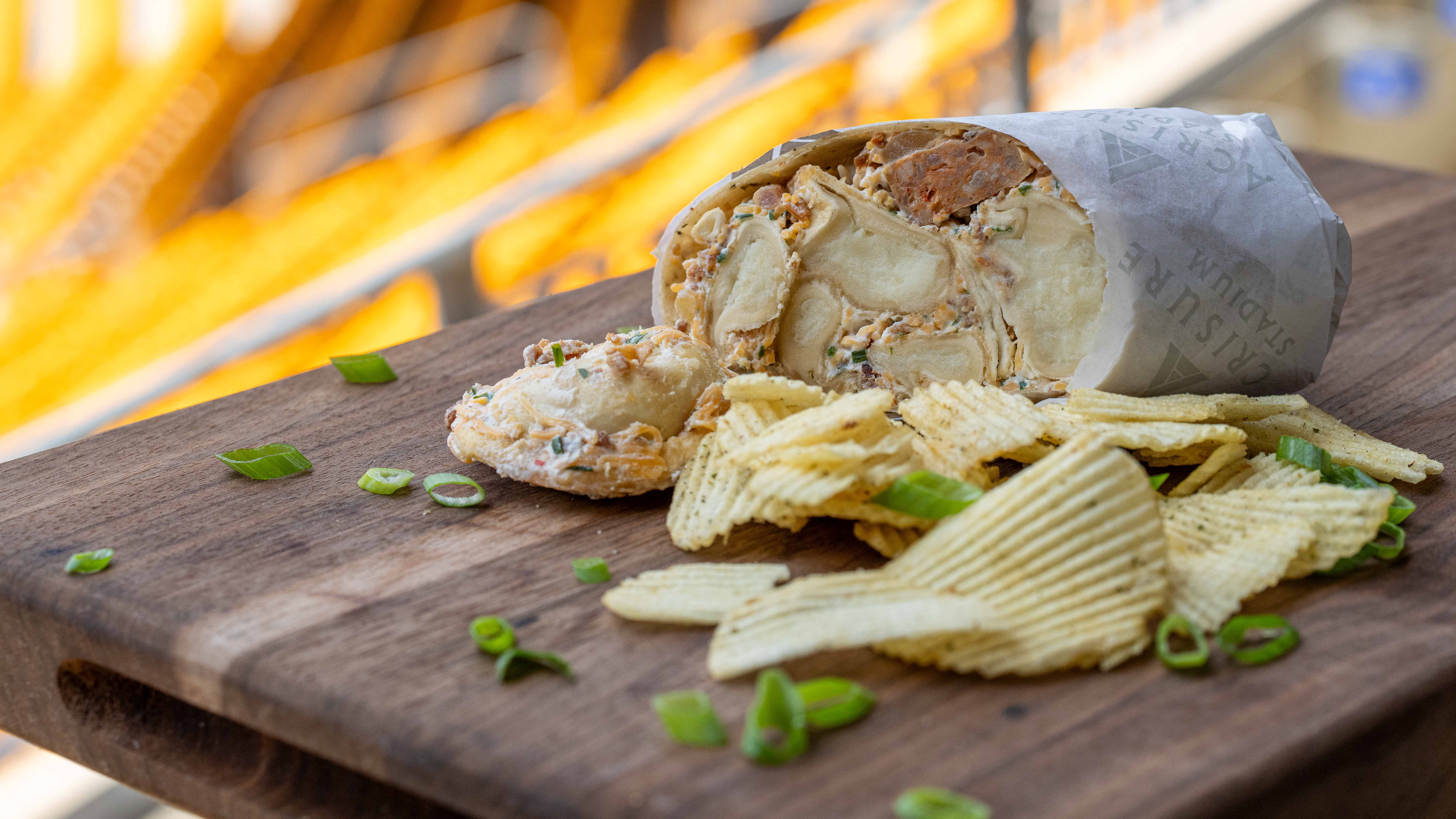 Close-up of a sliced wrap filled with pierogies and kielbasa on a wooden board, with ridged potato chips and scattered green onions; warm, blurred yellow background.