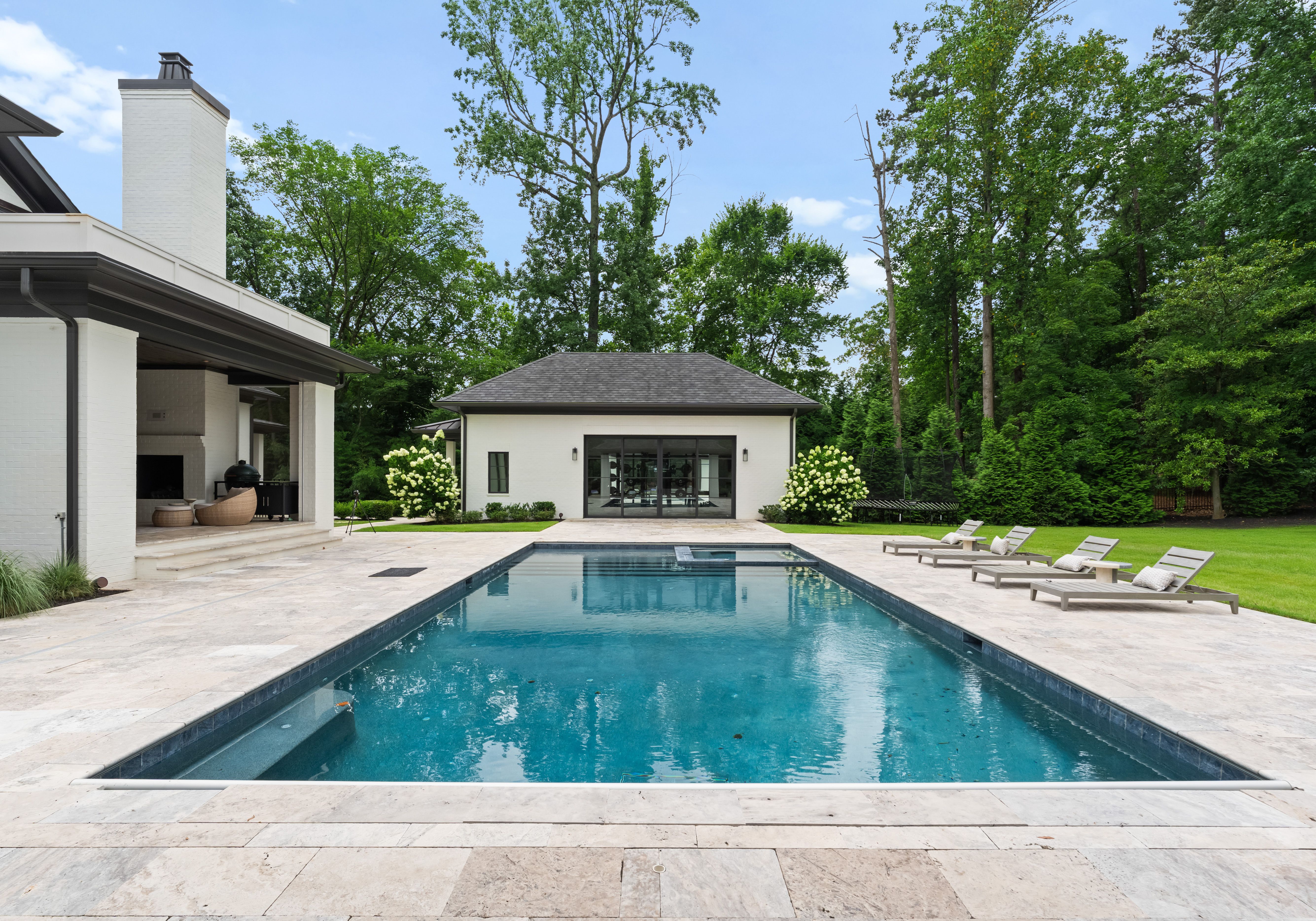Modern backyard with a rectangular blue swimming pool, beige stone deck, four beige lounge chairs with pillows, white buildings, green lawn, and tall trees under a blue sky.