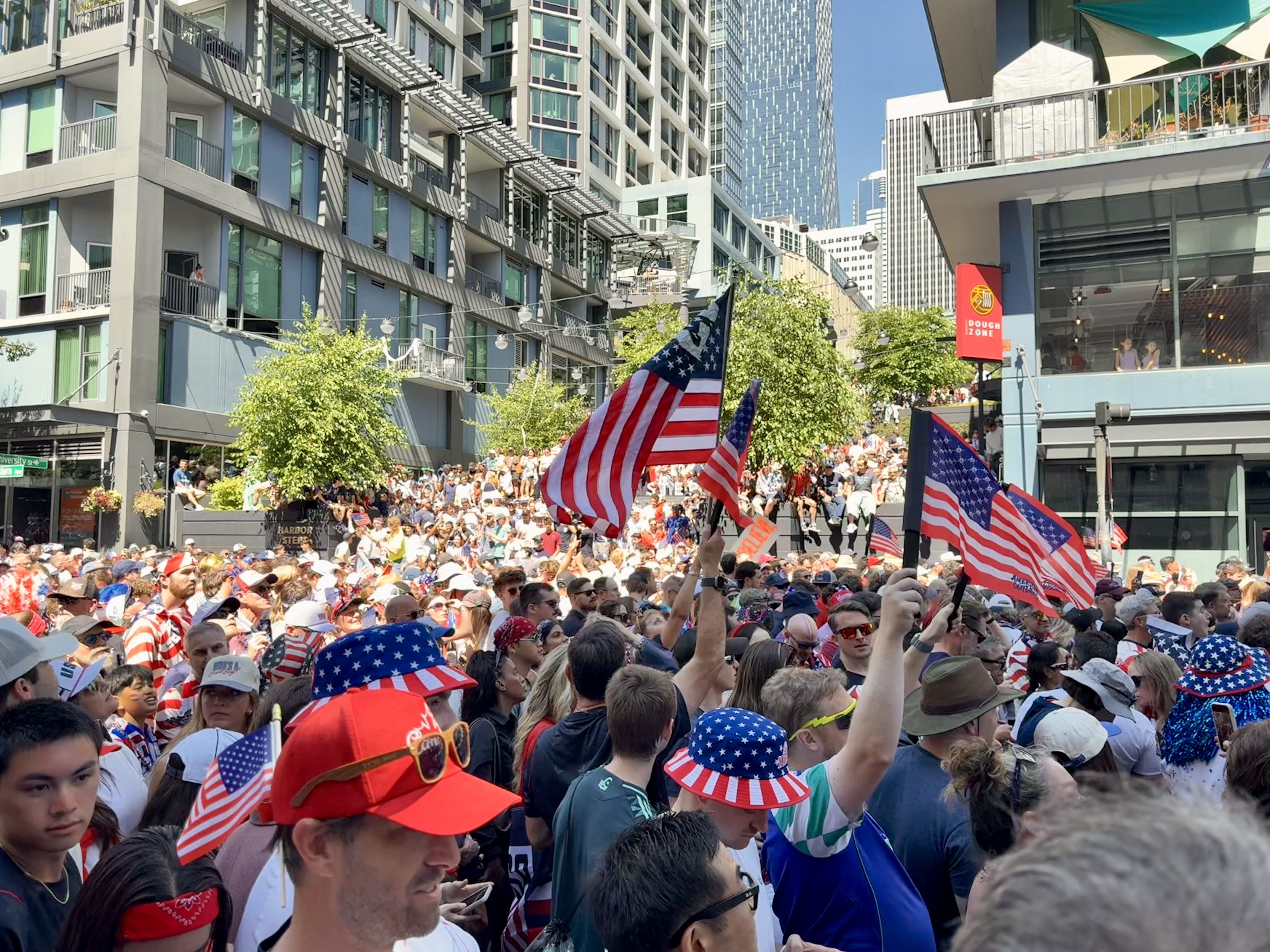 Large crowd fills a city street parade, waving red, white, and blue American flags. Modern glass-and-concrete buildings line both sides as spectators watch from balconies.