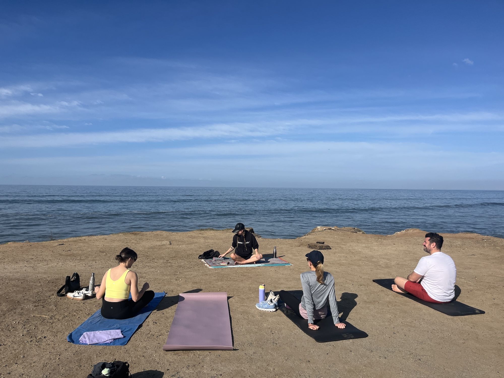 Four people sitting on yoga mats on a sandy cliff overlooking a calm blue ocean under a bright, partly cloudy sky.