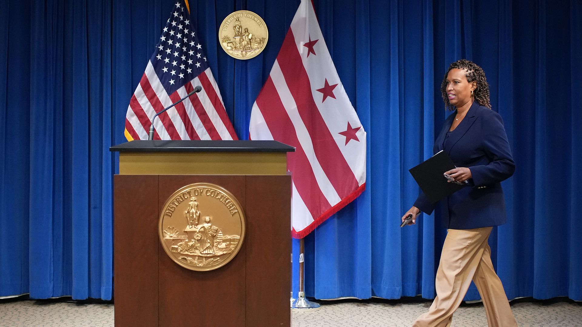 Mayor Muriel Bowser walks up to a podium at a city hall press briefing room, with the U.S. and D.C. flags behind