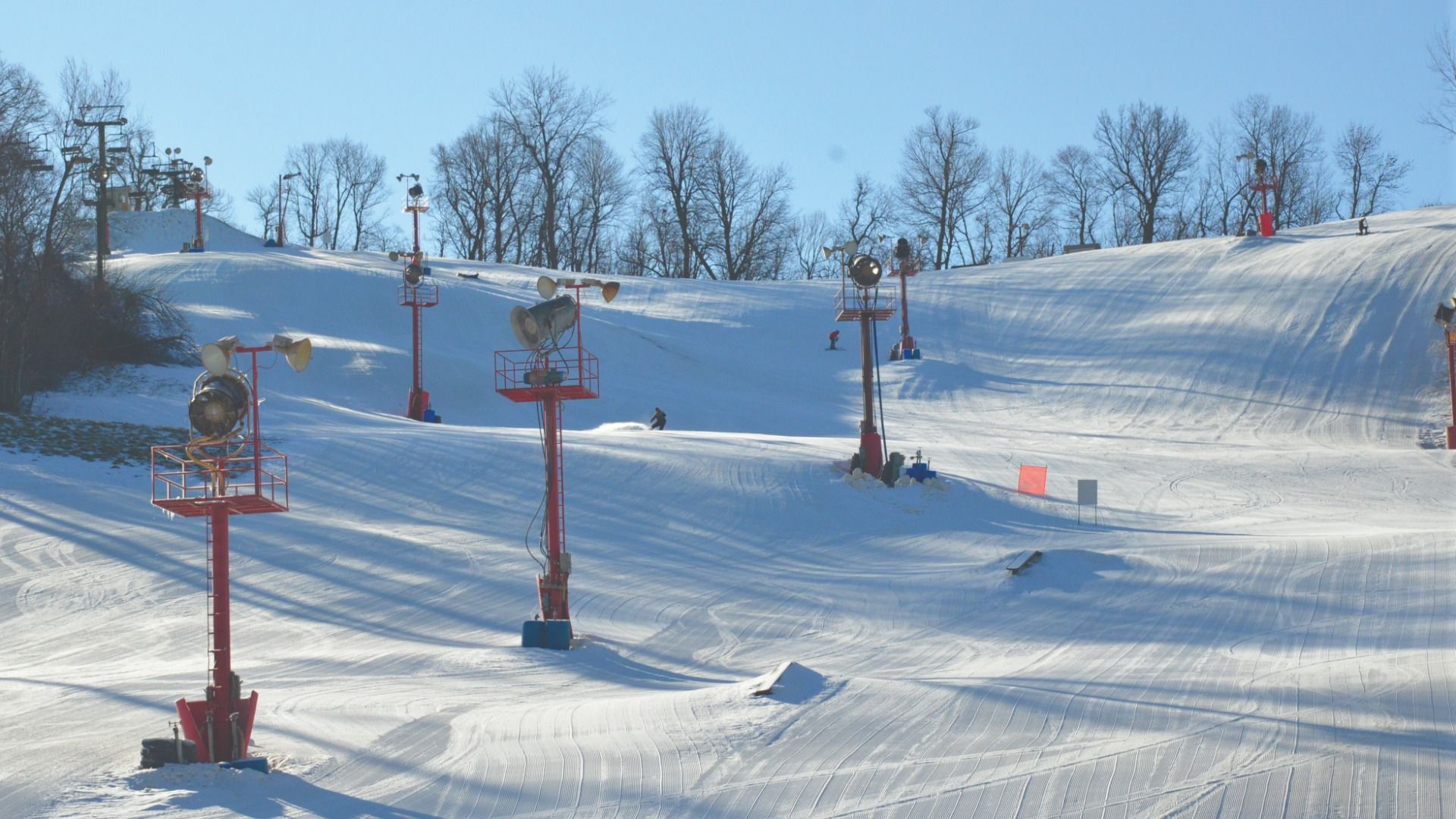 Bright sunny ski slope with fresh groomed snow, lined by tall red ski lift poles and leafless trees at Snow Creek in Missouri. A few skiers are descending the hill under a clear blue sky.