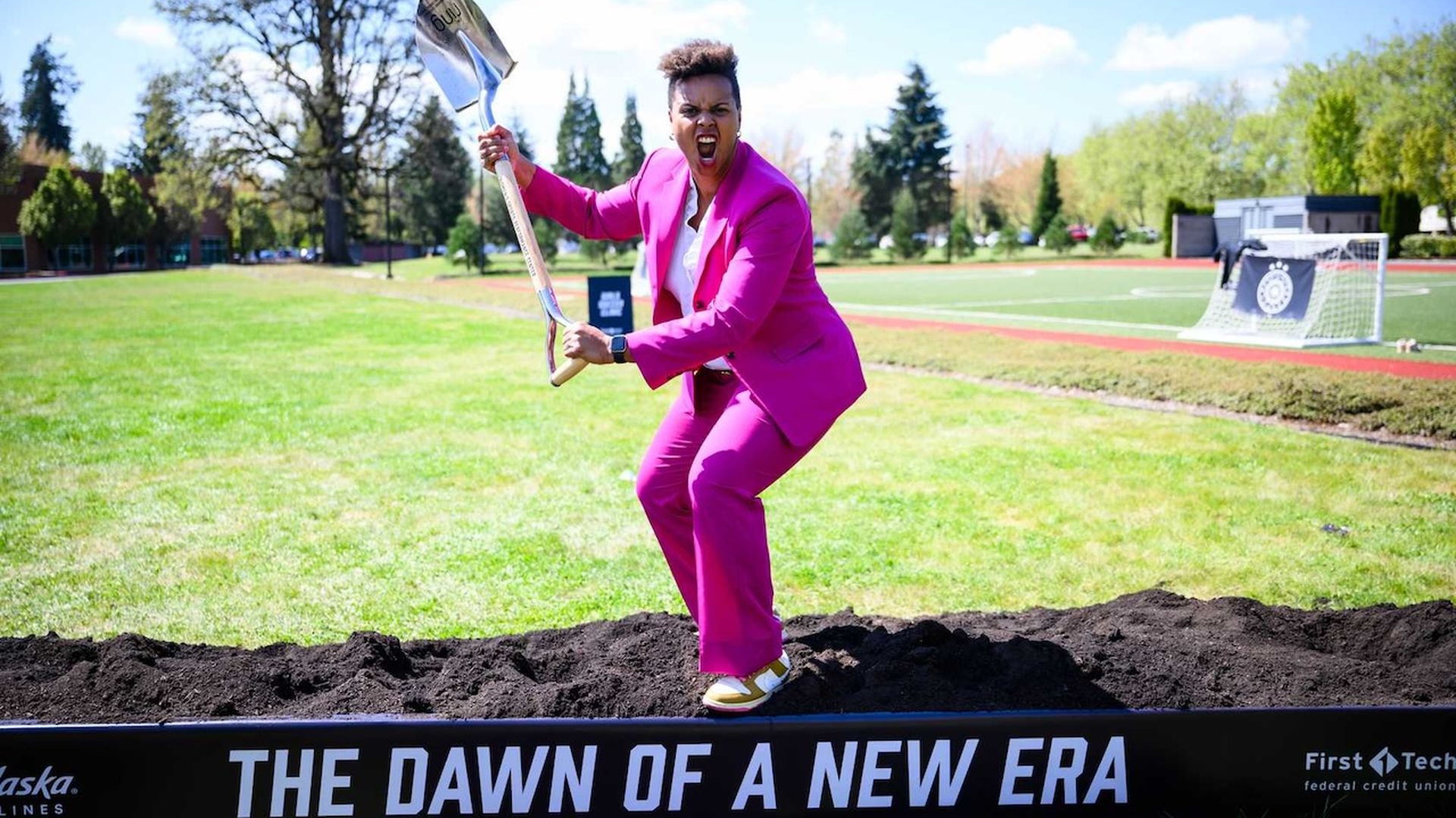 Person in a bright pink suit strikes a powerful pose with a shovel over fresh dirt at a groundbreaking event, standing on a platform labeled "The Dawn of a New Era" on a sunny day near a sports field.