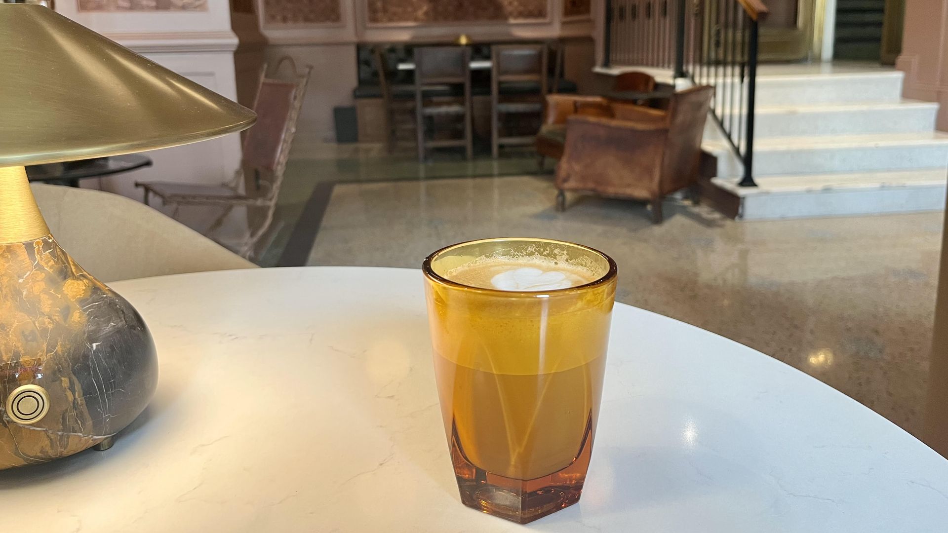 A latte in an orange-tinted glass sits on a white table next to a brown and gold lamp.