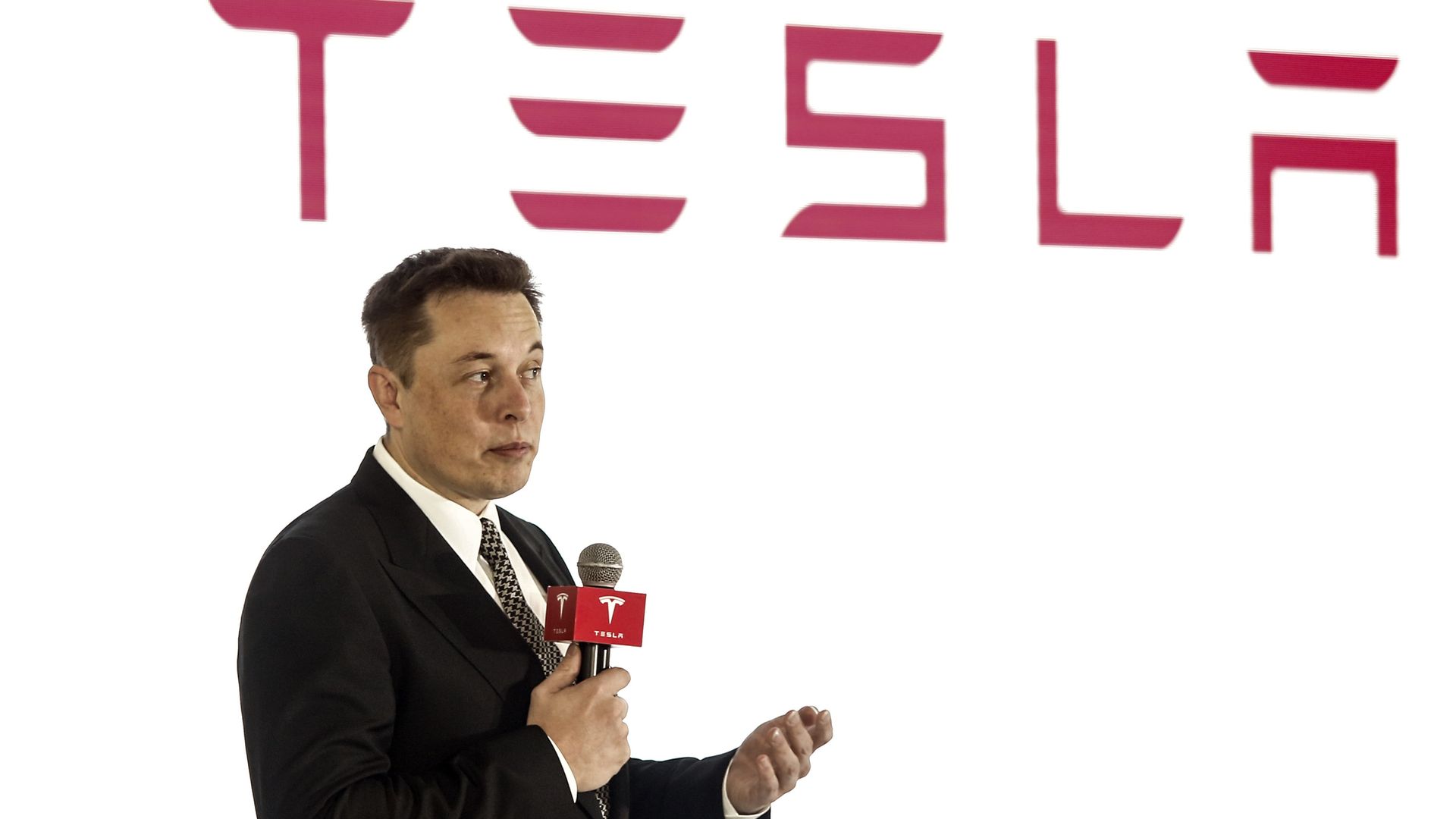 Elon mUsk in a suit on a stage with a microphone.