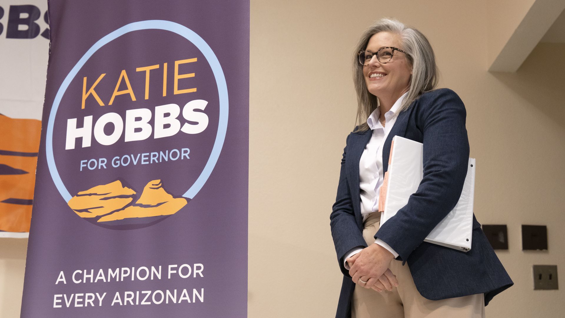 A smiling woman in glasses wearing a dark blue or black blazer over a white shirt and khaki pants stands next to vertical banner that reads "KATIE HOBBS FOR GOVERNOR" in a circle with a mountain logo, above the words "A CHAMPION FOR EVERY ARIZONAN".