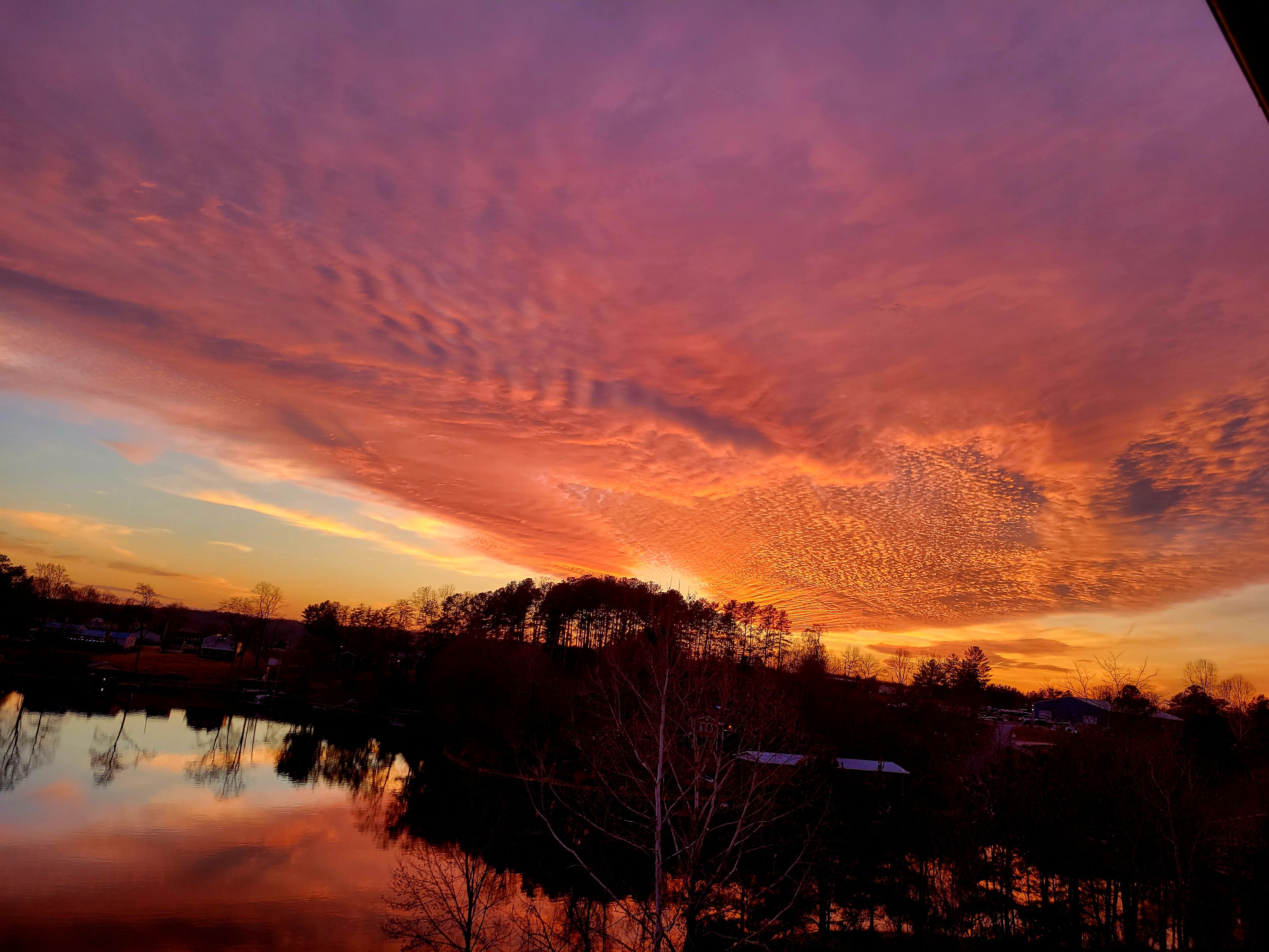 Vivid sunset with purple and orange clouds over a lake, reflecting the sky. Silhouetted trees and houses line the horizon under the dramatic colorful sky.