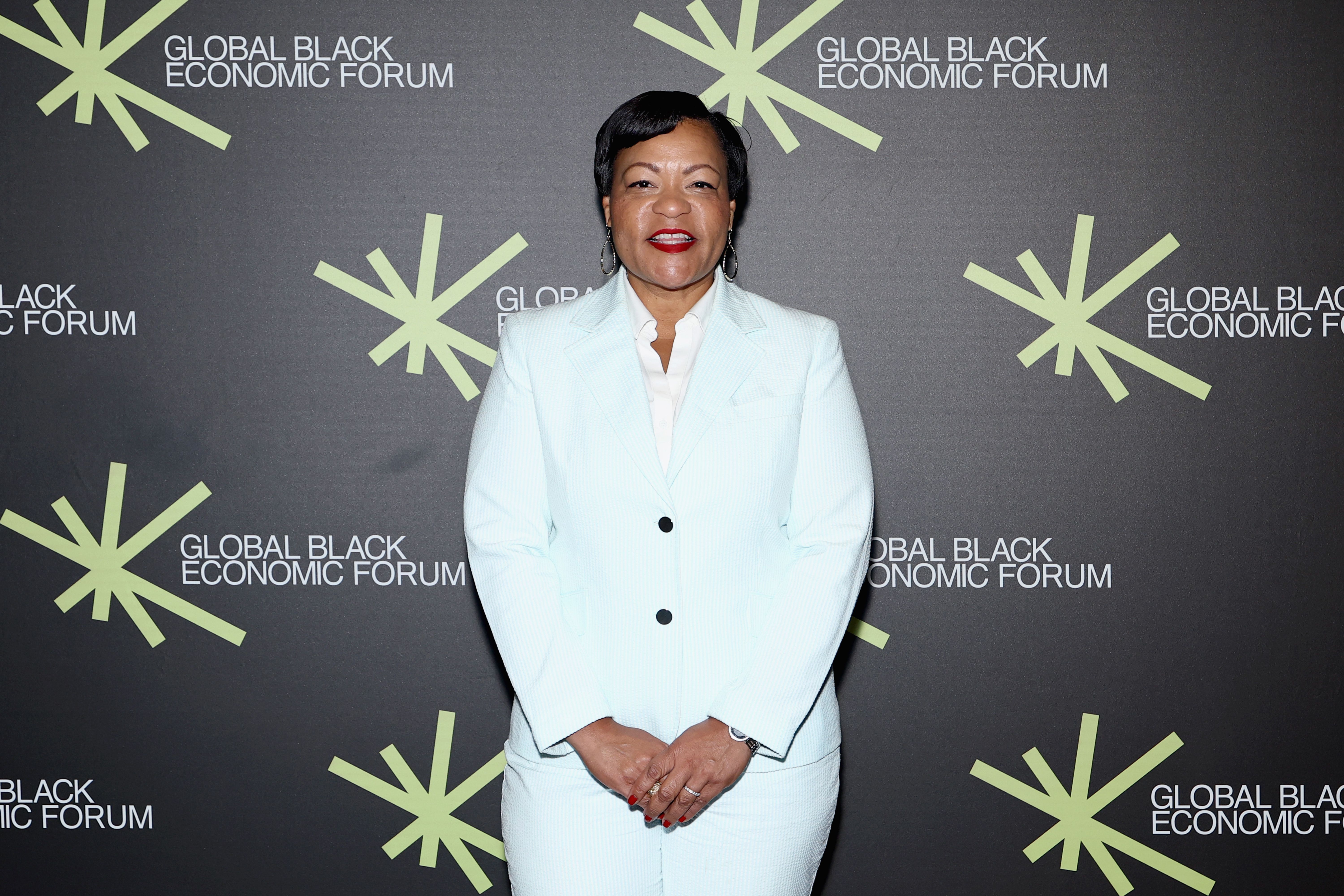 Woman in a light blue suit and white shirt stands in front of a black backdrop with green starbursts and "Global Black Economic Forum" text multiple times.