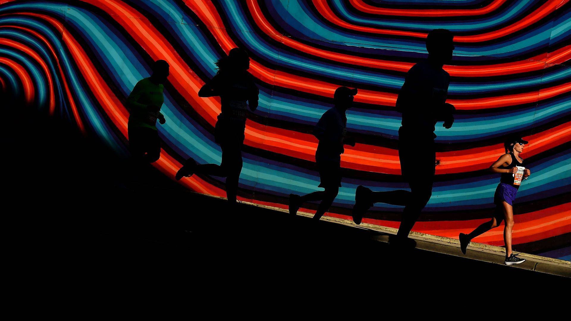 A group of people, some in shadows, run past a mural of colored waves. 