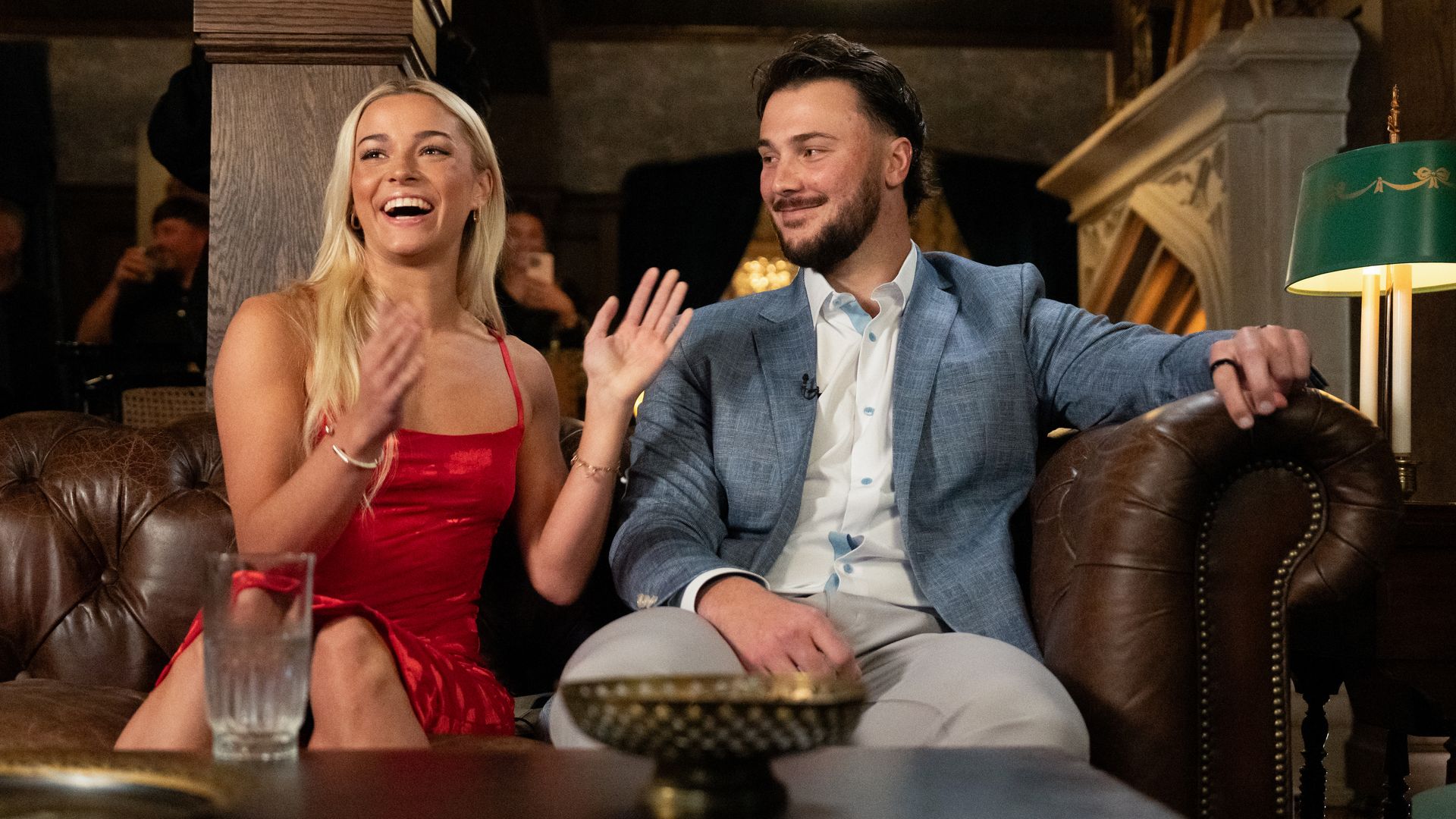 Livvy Dunne and Paul Skenes sit on a brown leather sofa. Dunne is mid-clap as she looks over the photographer's shoulder, and Skenes looks at her.
