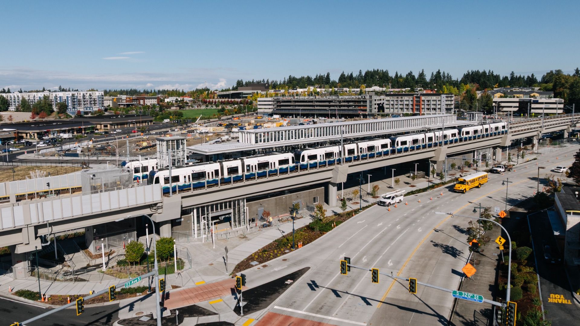 Elevated light rail train on a concrete bridge above a wide road intersection with traffic lights, sidewalks, a yellow school bus, and buildings in the background under a clear blue sky.