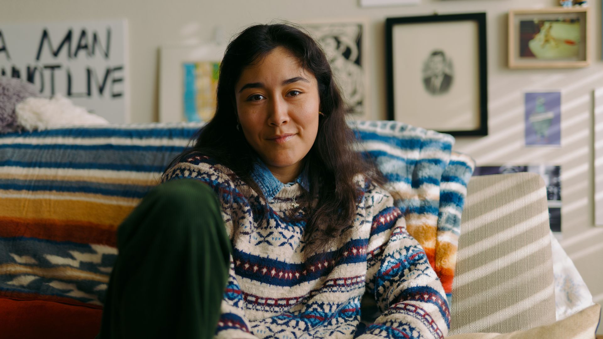 A young woman with dark hair sits on a striped blue, white, and orange blanket on a couch in a cozy room; framed art and photos hang on the wall behind her.