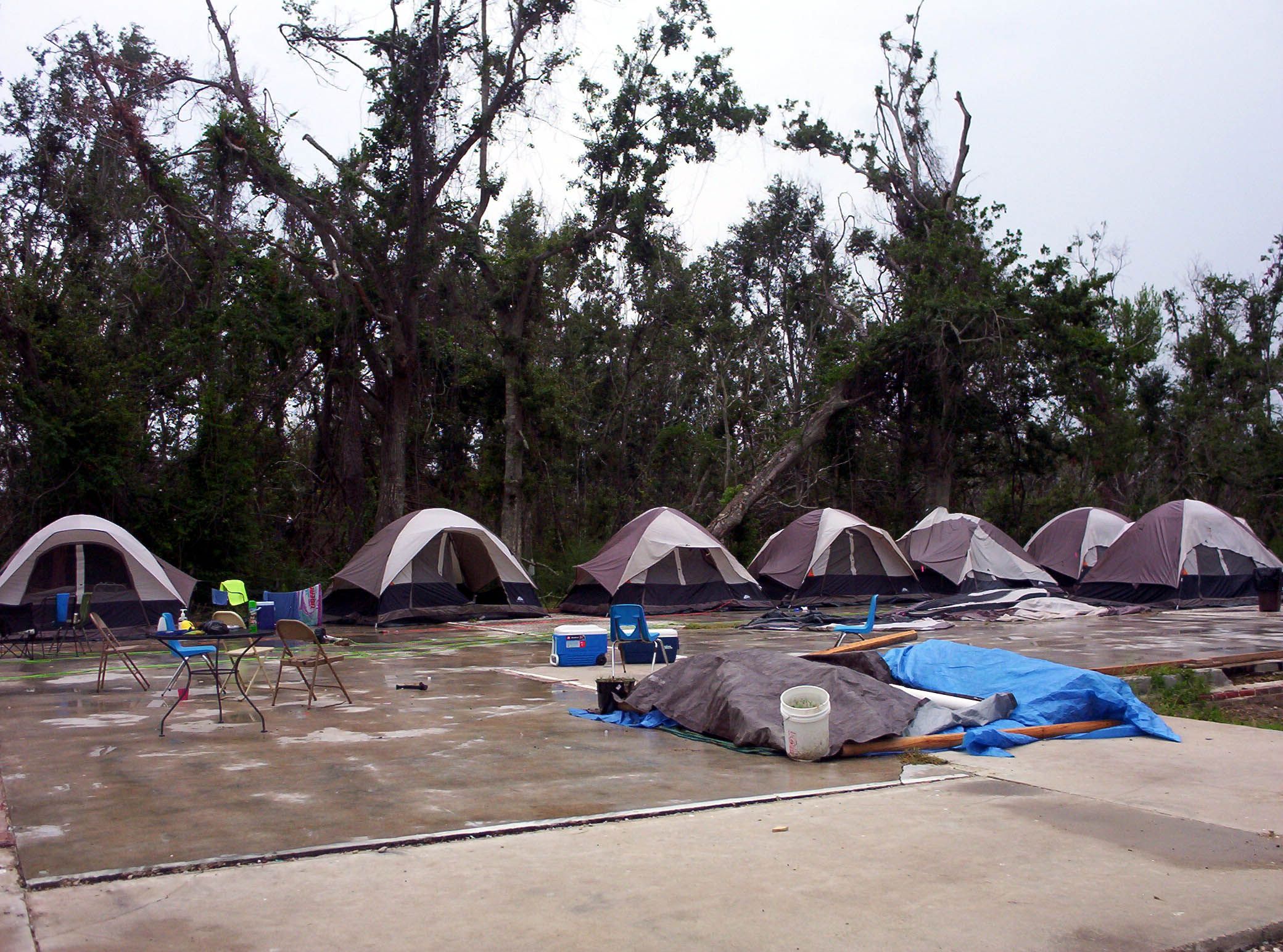 Multiple small tents set up on a concrete slab with scattered chairs and covered items, surrounded by tall trees under a cloudy sky.