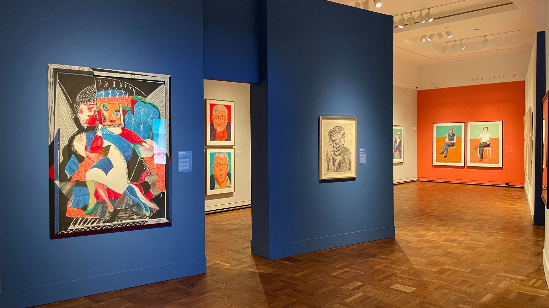 Art gallery with bright blue and orange walls displaying colorful modern and abstract portraits, including one with distorted facial features and another of seated figures on orange background.