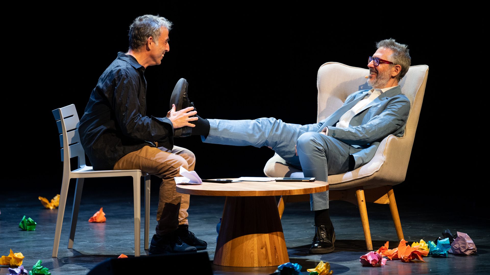 Etgar Keret is sitting facing Ira Glass, who has his right leg up and Keret is holding Glass's foot. They are on a stage with a wood table between them and colorful paper on the ground.