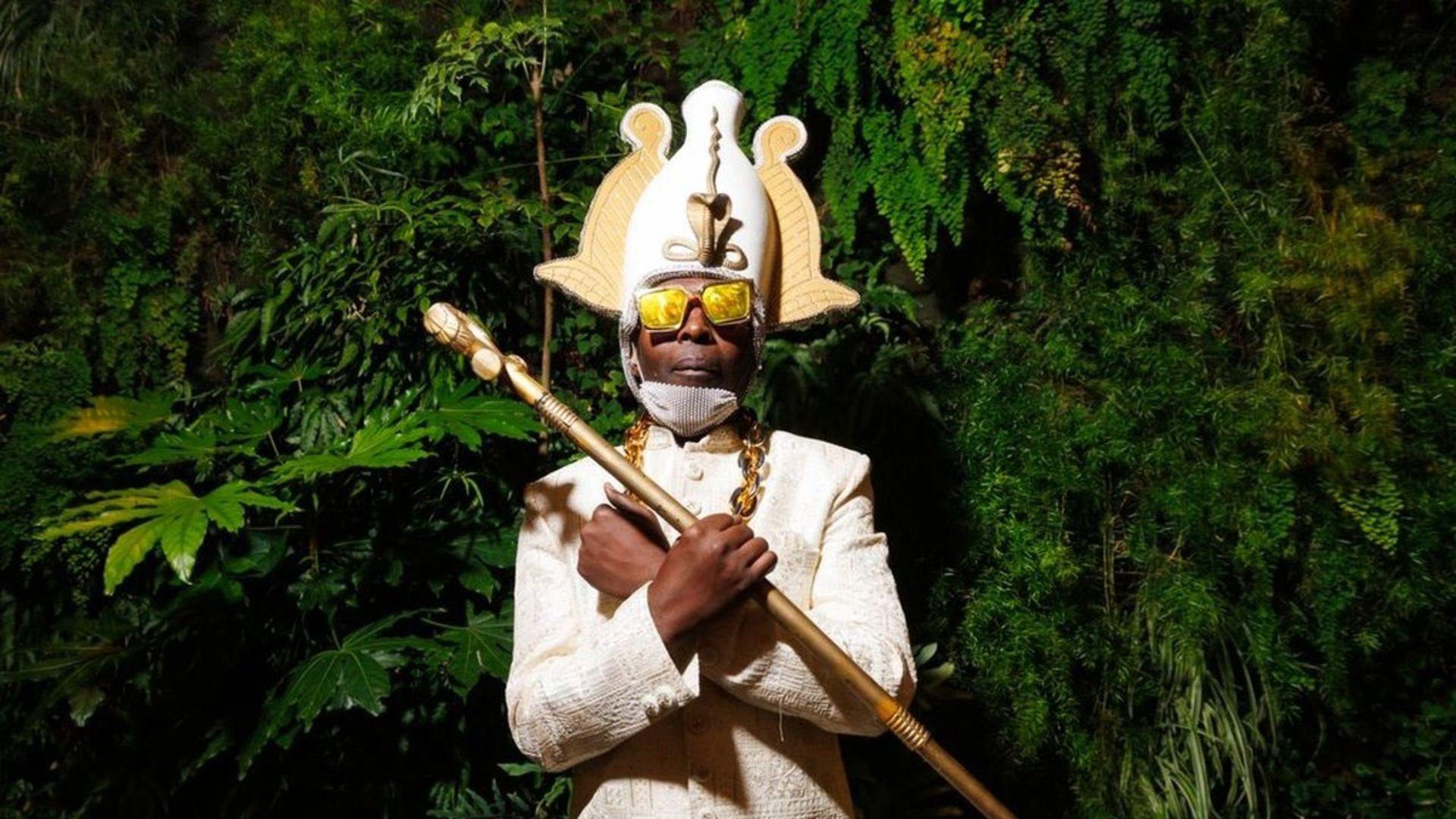 A person in an ornate cream suit wears a large gold headdress and yellow sunglasses, standing before dense green foliage while holding a long gold staff across the chest.