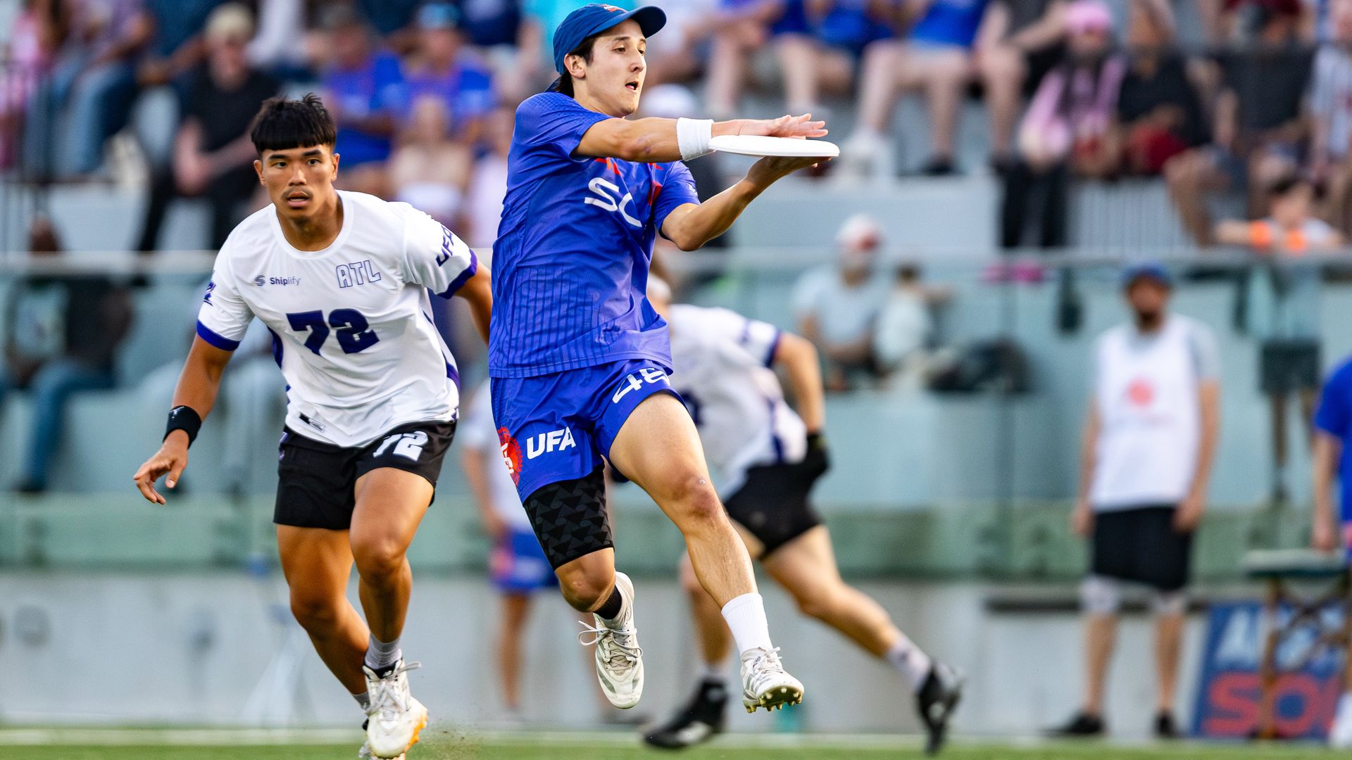 Action shot of a blue-uniform player throwing a frisbee as a white-uniform opponent chases; green field in foreground and a blurred stadium crowd in the background.