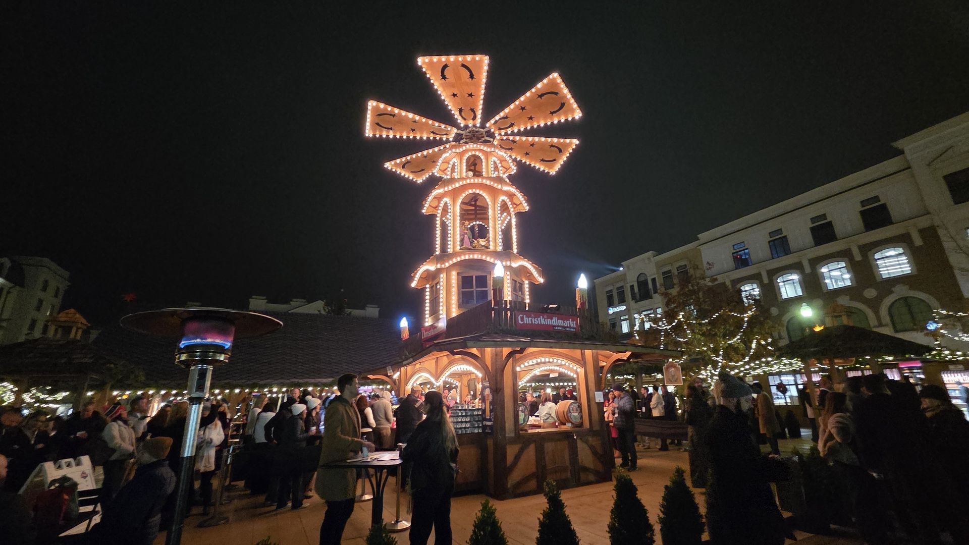 Night scene at a Christmas market with a large illuminated wooden tower decorated with lights and star and moon patterns, surrounded by people and string-lit trees.