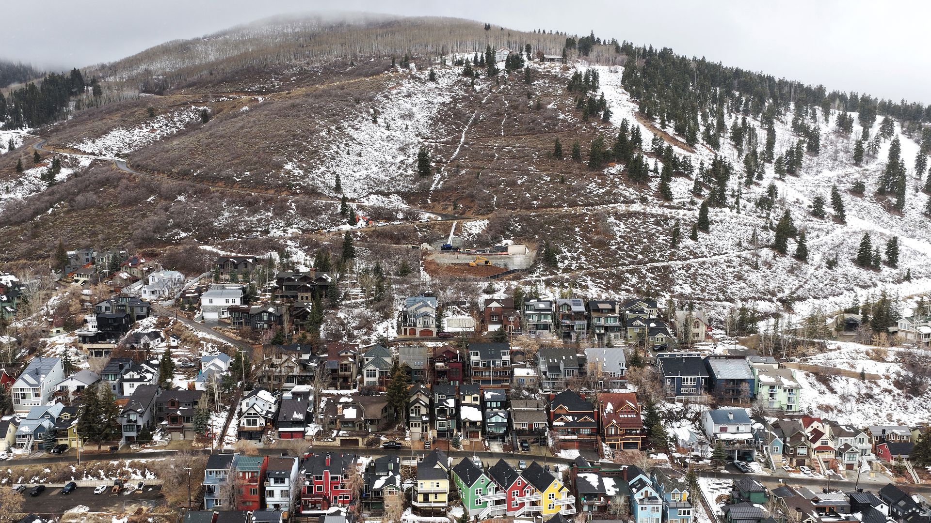 A snowy hillside town with many colorful houses including red, green, yellow, and blue, surrounded by leafless trees and a partially snow-covered mountain in the background under gray sky.