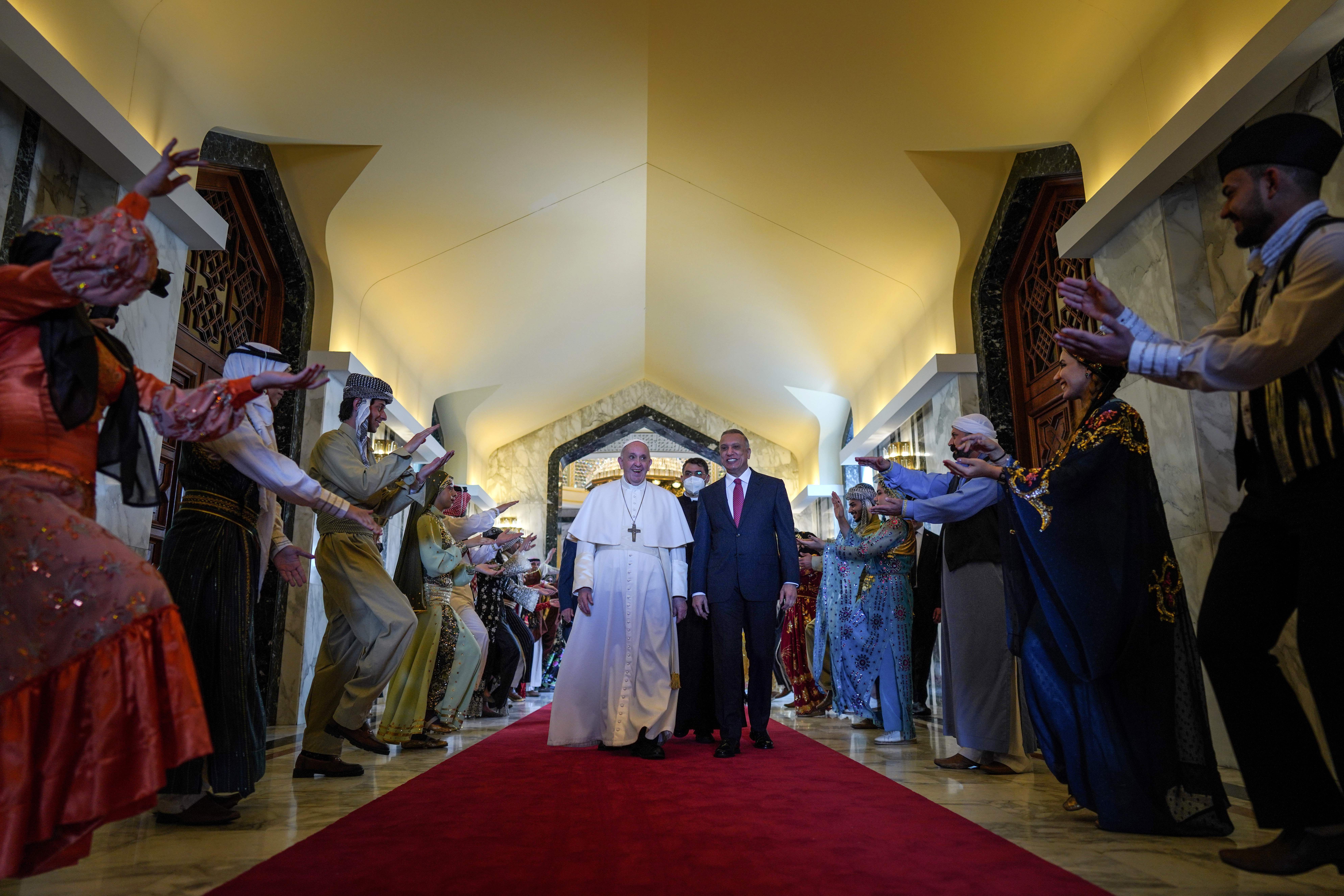 Photo of Pope Francis walking next to Iraq's Prime Minister Mustafa al-Kadhemi in a hallways surrounded by performers.