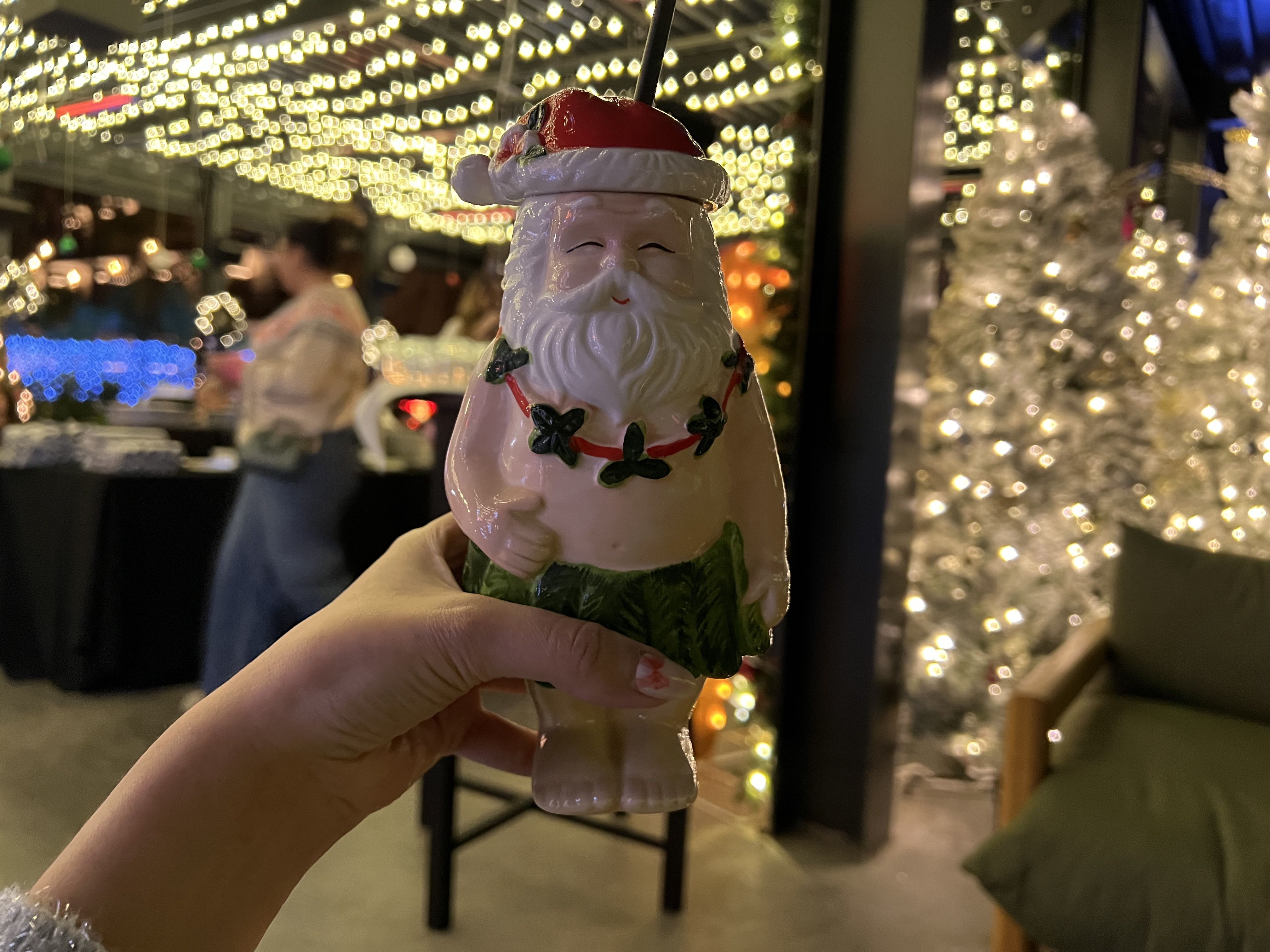 Hand holding a Santa-shaped cup wearing a red hat and green skirt, with string of green leaves around torso, against blurred background of warm twinkle lights and decorated white Christmas trees.