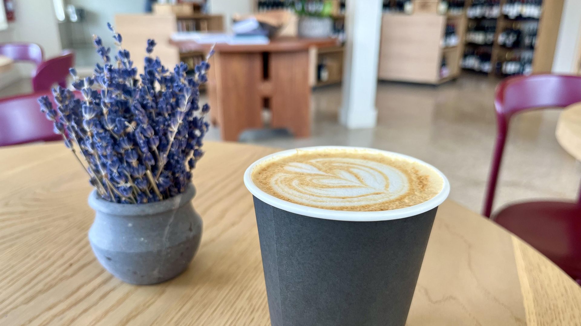 A latte in a to-go cup, with a bunch of lavender sitting on the table in the background, and a wine shop farther in the background.