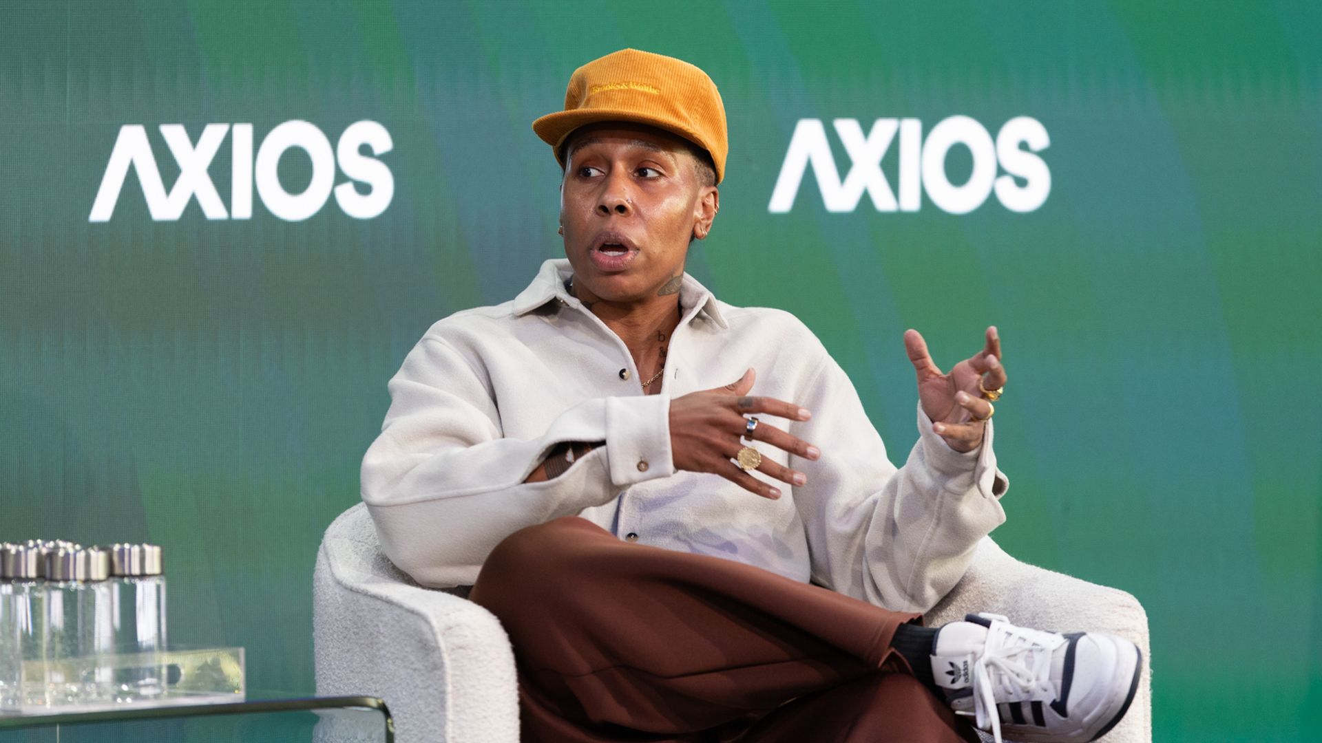 A person in an orange cap, beige shirt, brown pants and white sneakers sits in a gray chair on a stage with a green Axios backdrop, gesturing with hands during a discussion.