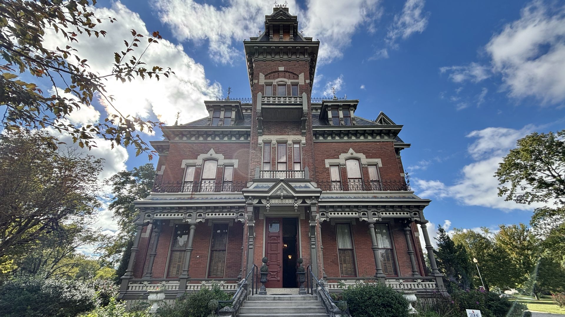 Red brick Victorian mansion with a tall central tower, decorative trim, arched windows, and wrought-iron railings. Front steps rise to carved doors, surrounded by trees under a blue sky.