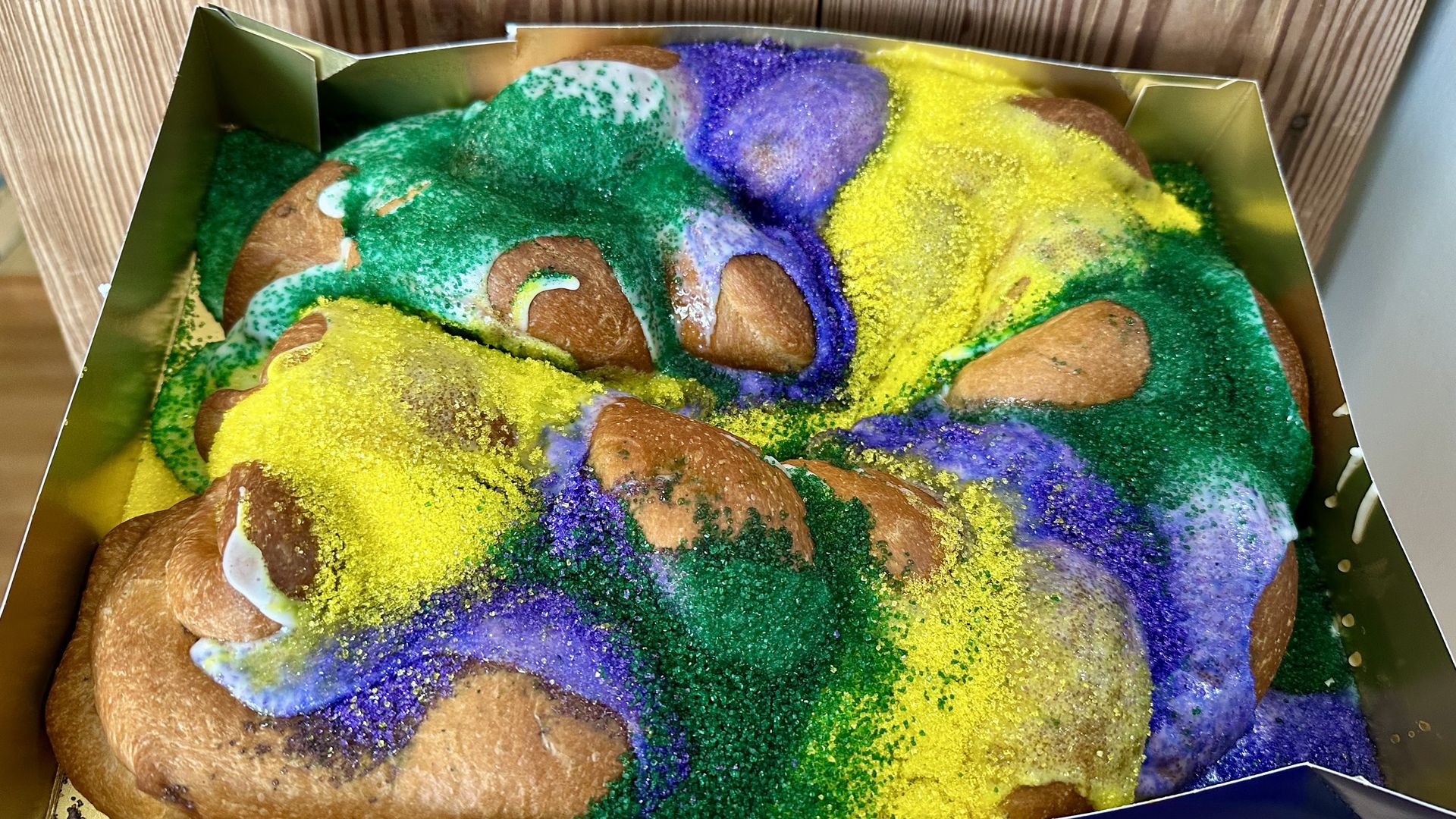 Close-up of a King Cake with purple, green, and yellow sugar sprinkles and icing, placed in a gold-colored box on a wooden surface background.