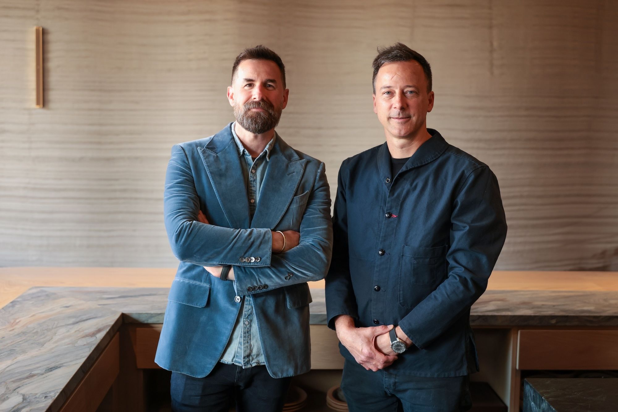A man in blue, velvet sport coat and a chef in a black shirt stand inside their restaurant. 