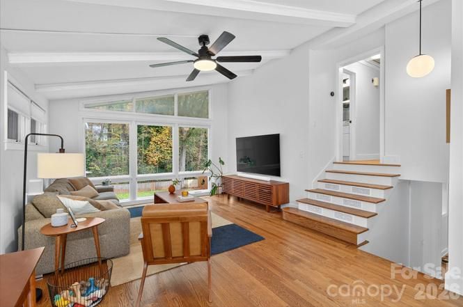Bright living room with large windows showing autumn trees, wooden floors, gray sofa, wooden armchair, coffee table, ceiling fan, TV on wooden stand, and stairs leading up on the right.