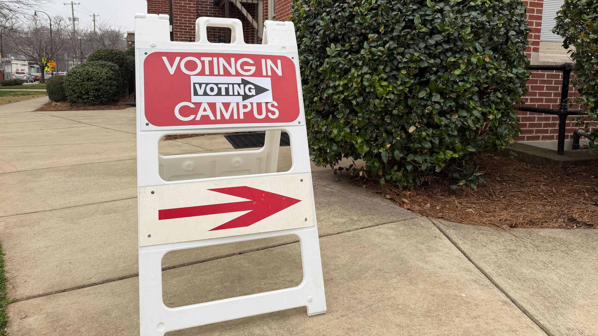 White sign on sidewalk shows red text "VOTING IN CAMPUS" with black arrow labeled "VOTING" pointing right, directing to a brick building with bushes on a cloudy day.