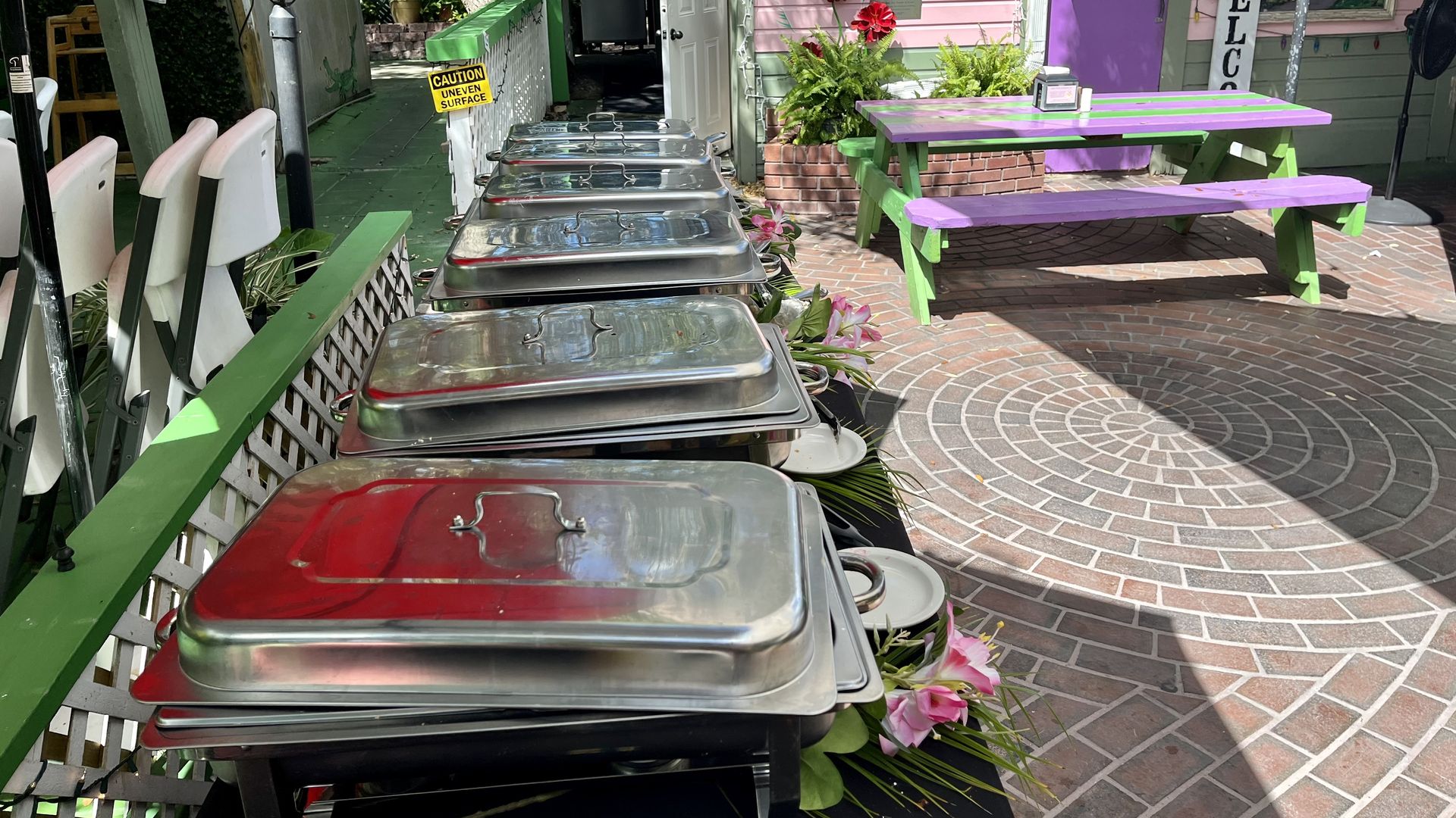 A table with five silver chafers in a brick courtyard near a picnic table painted purple and green.