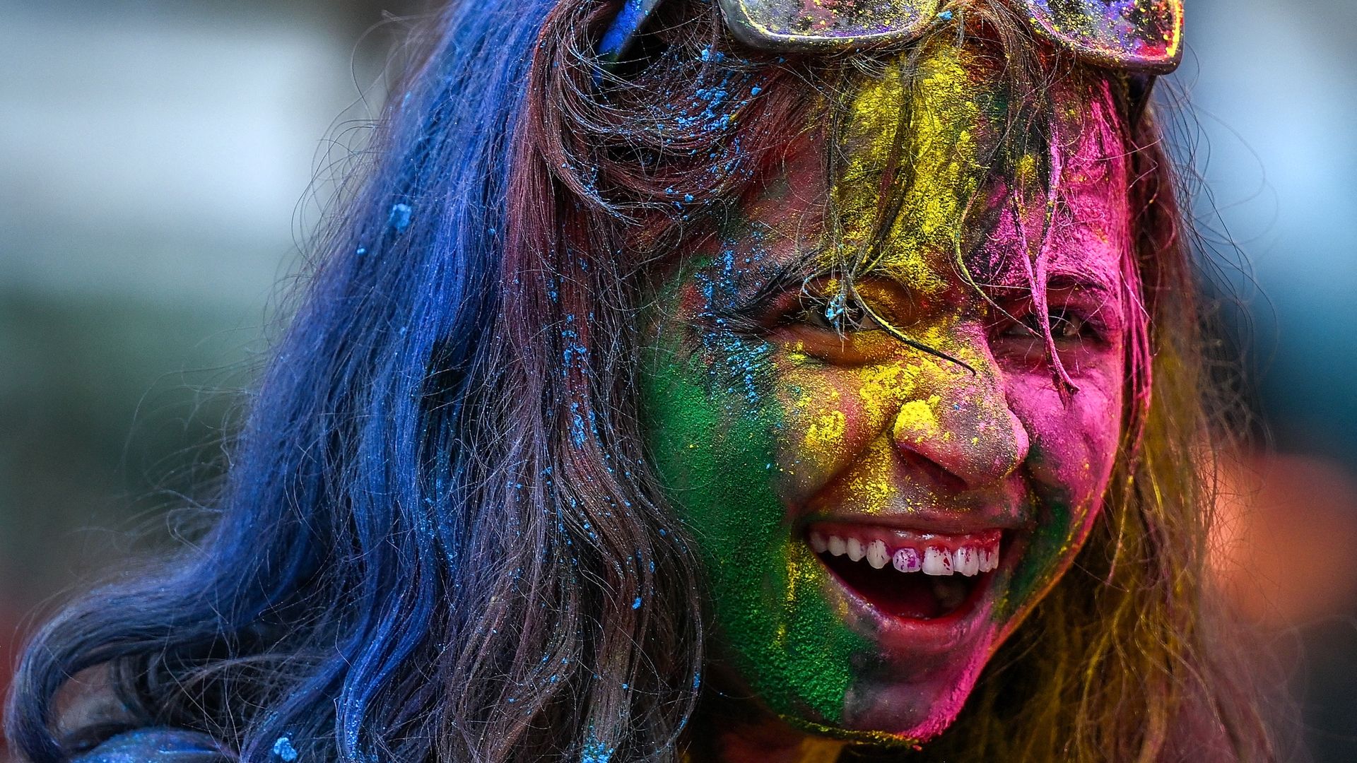 Image shows a woman smiling and covered in blue, green, yellow and pink powder.