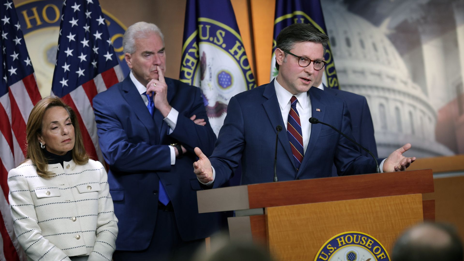 Man in blue suit and glasses speaking at U.S. House of Representatives podium, flanked by two people, one in white jacket and one in blue suit, with American flags in background.