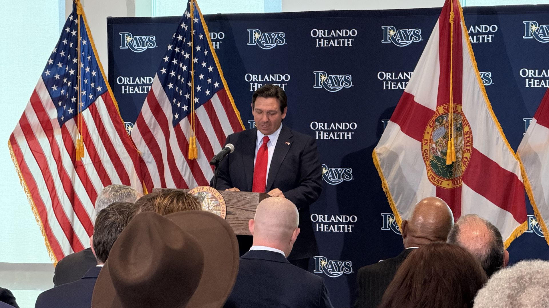 A man in a dark suit and red tie speaks at a podium with the Florida state seal, flanked by U.S. and Florida flags, with a backdrop featuring "Rays" and "Orlando Health" logos.