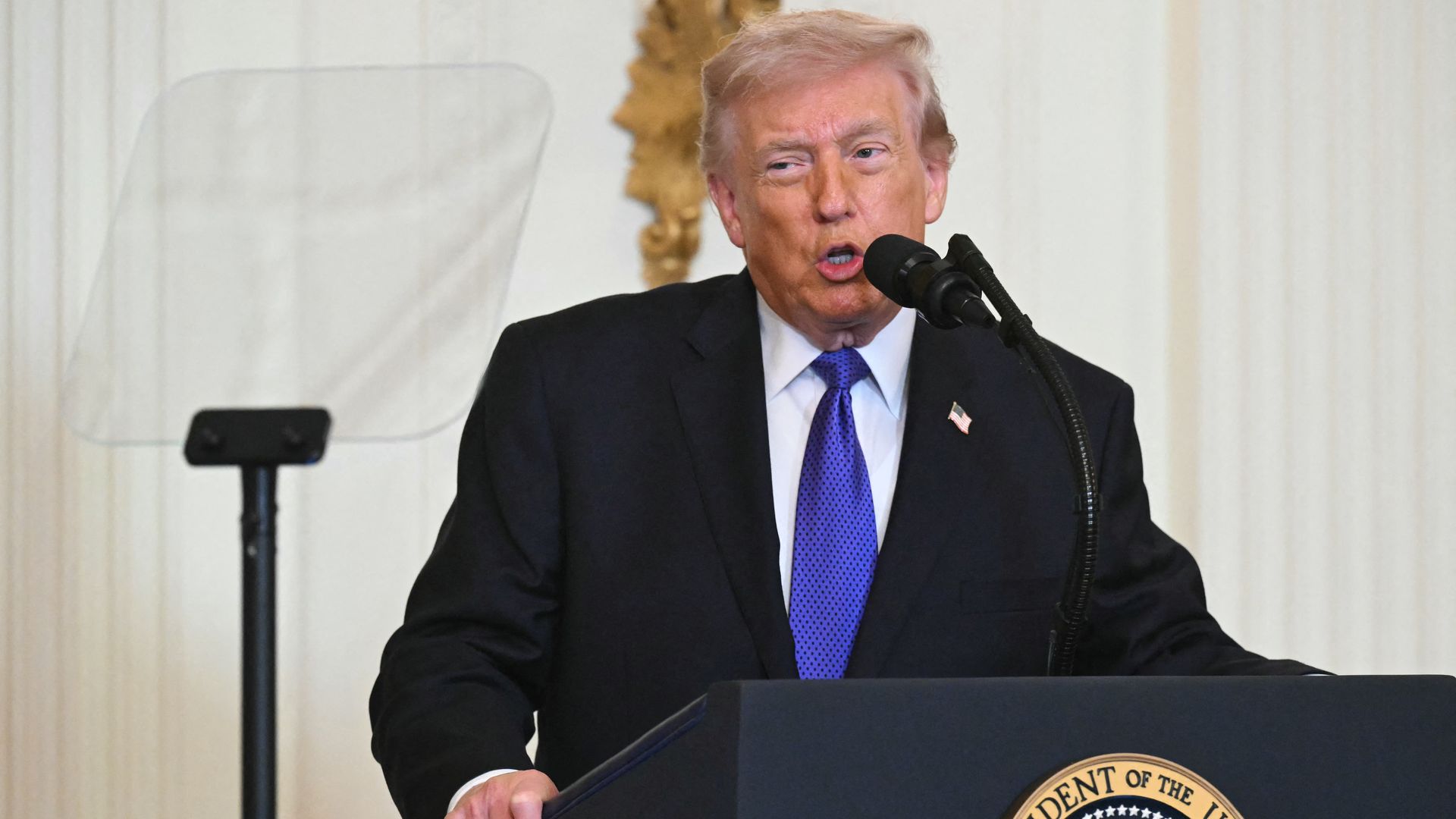 President Trump, with graying strawberry blond hair and a blue tie. speaks at a podium with the presidential seal, wearing a black suit and standing next to a clear teleprompter screen in a formal setting.