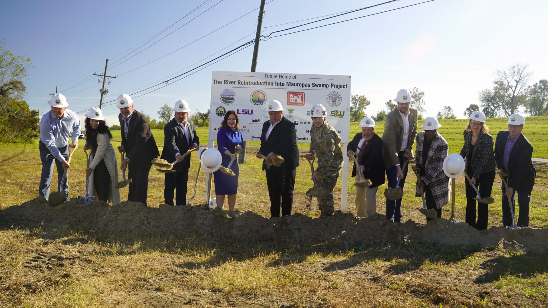 A line of a dozen people wearing white hard hats turn over dirt in a ceremonial groundbreaking with signage behind them.