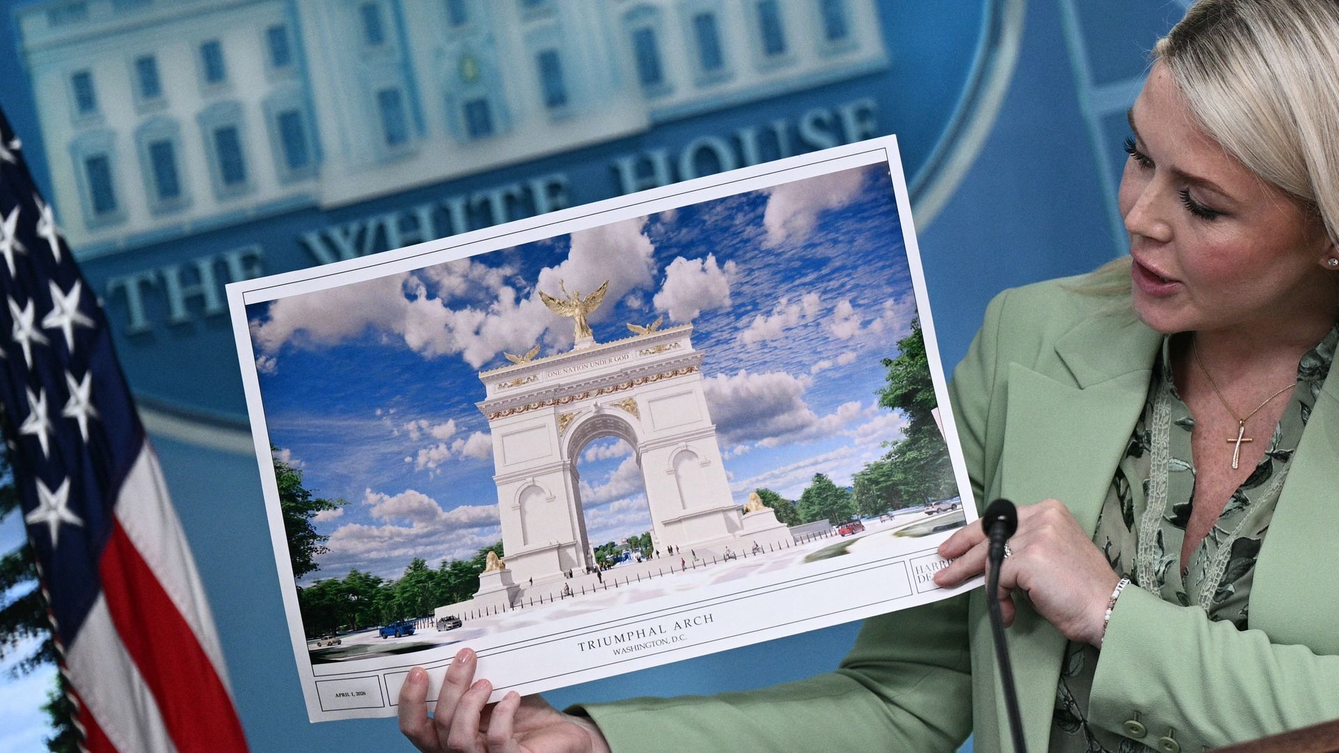 A blonde woman in a pale green blazer holds a large poster depicting the Arc de Triomphe in Washington, D.C., during a briefing, with an American flag and a blue backdrop behind her.