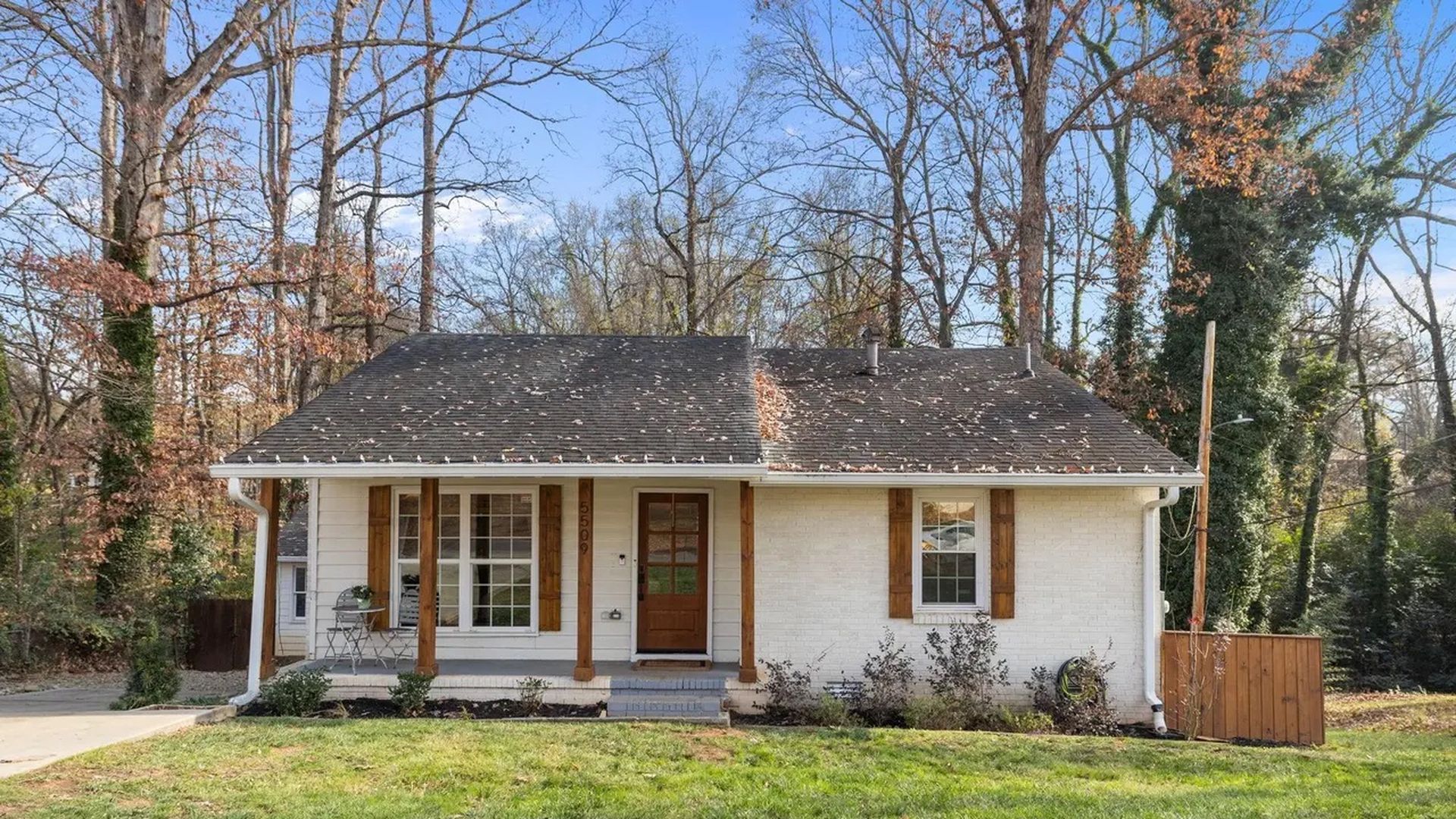 Single-story white brick house with dark shingled roof and wooden door, two windows with wooden shutters, small porch with two chairs, surrounded by leafless trees and green lawn.