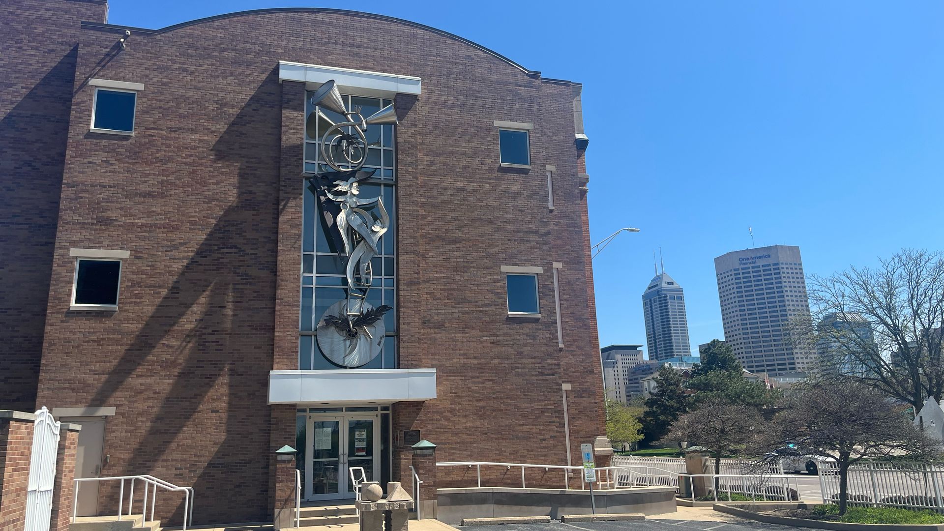 Brick building with a tall glass panel sculpture featuring a circular wheel and abstract metal figures; entrance with stairs, blue sky, and distant city skyline.