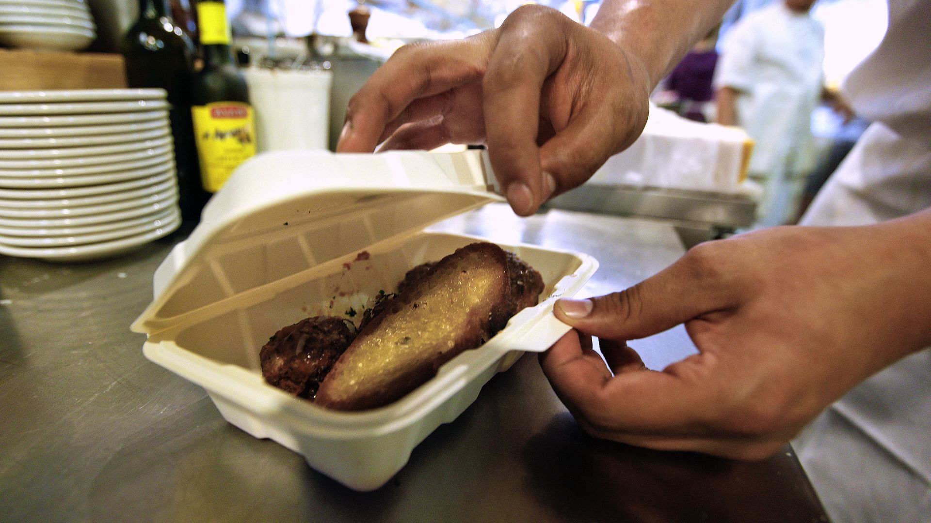 Two brown-skinned hands close a cream-colored takeout container of food