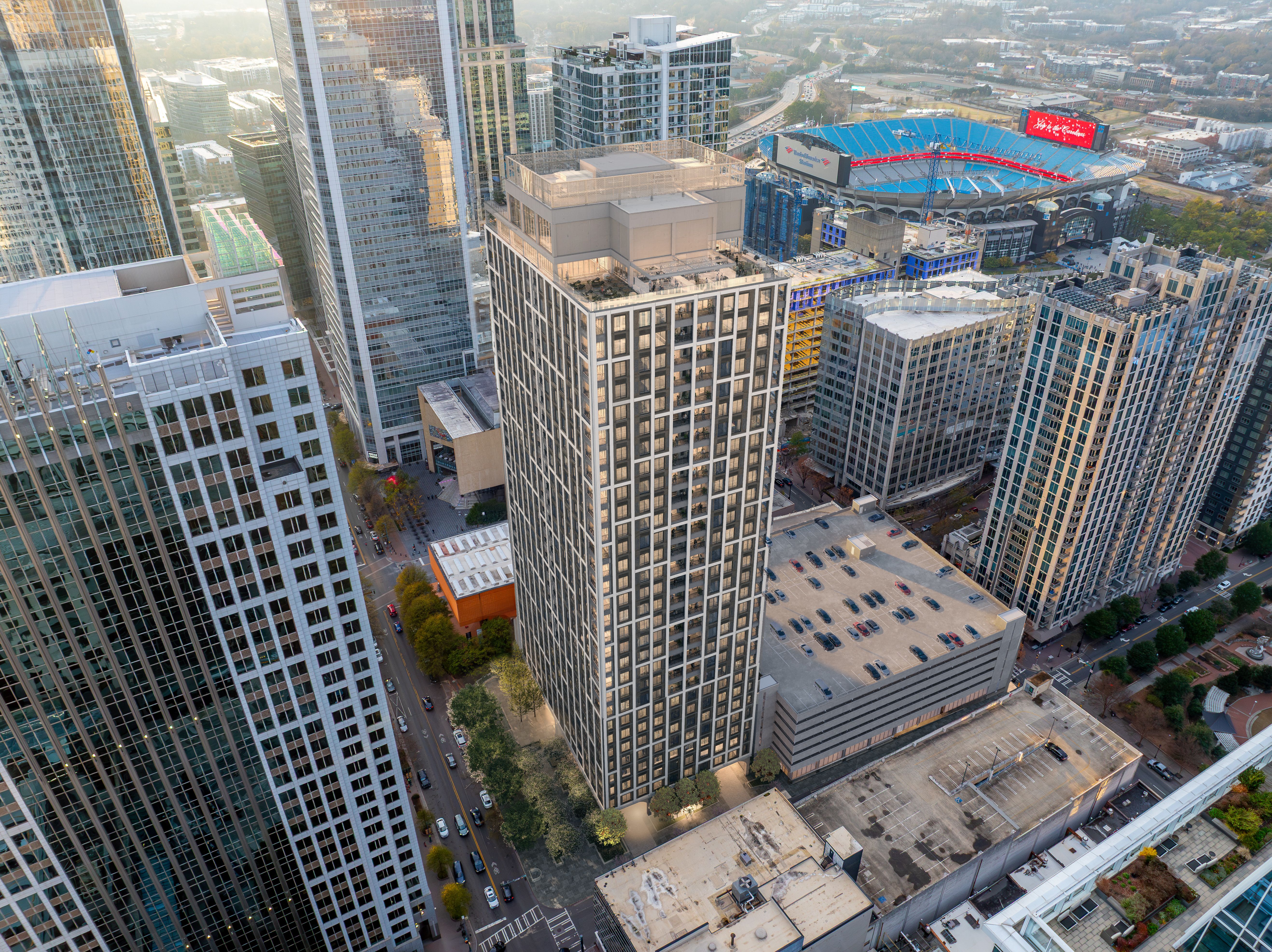 Aerial cityscape of tall glass skyscrapers; in the distance a large stadium with blue seating and a red video display, surrounded by roads, midrise buildings, and a parking lot.