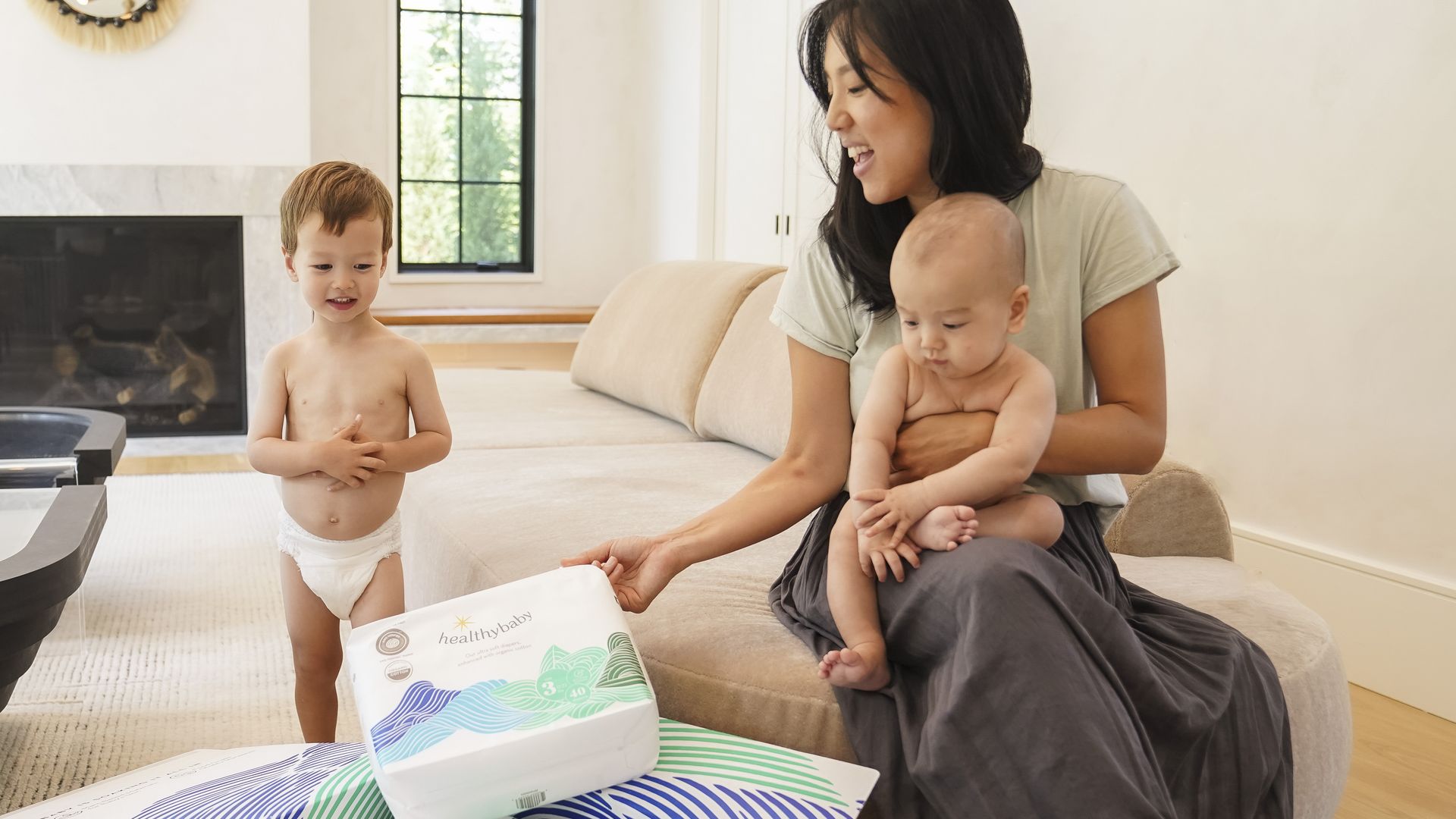 A mother with dark hair sits in a living room swathed in a blanket with her infant and toddler as she examines a package of Healthybaby diapers.