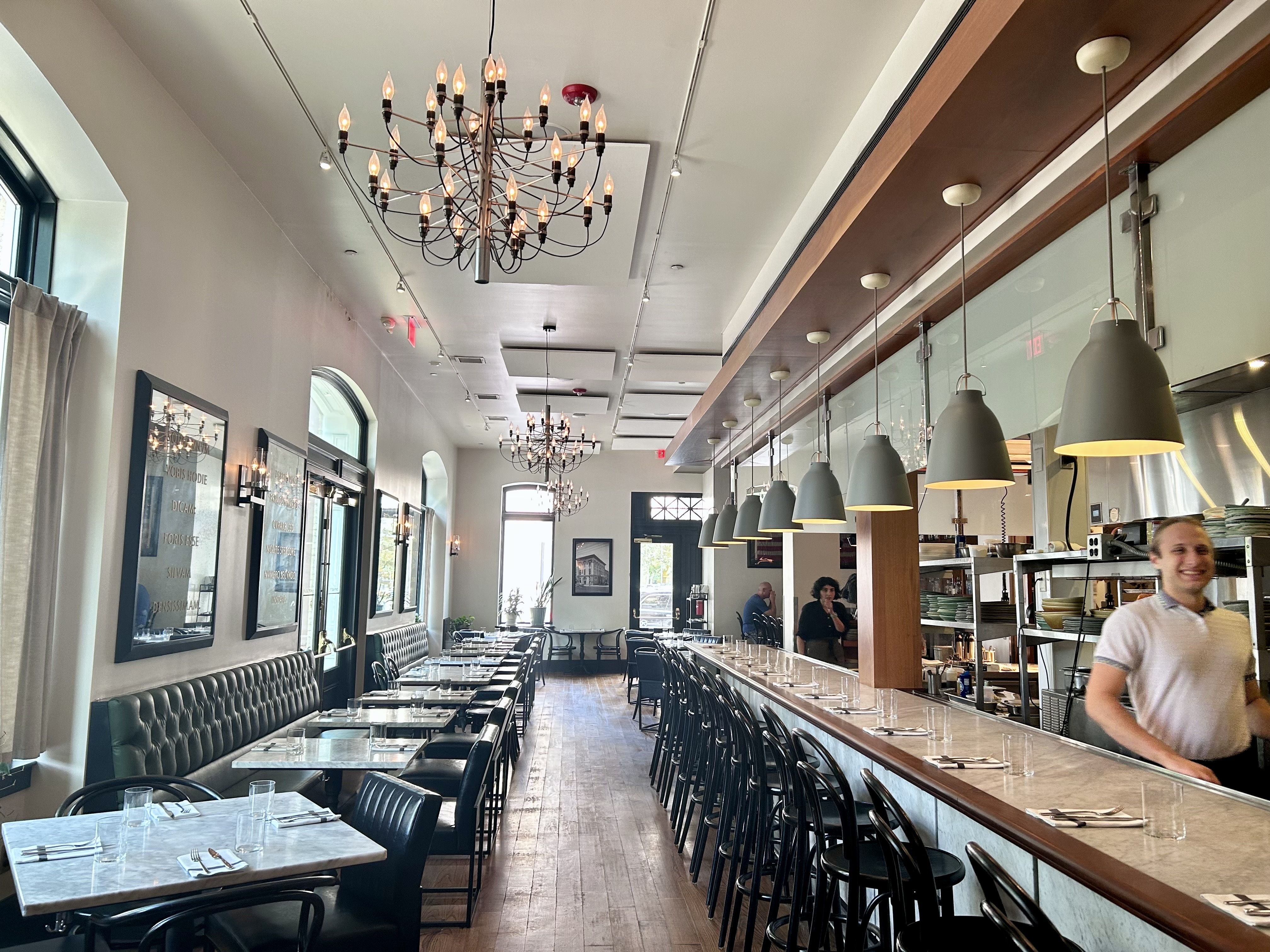 Bright modern restaurant interior with long marble bar, black stools, pendant lights, tufted bench seating, chandeliers, wooden floors, and a smiling server near the kitchen.