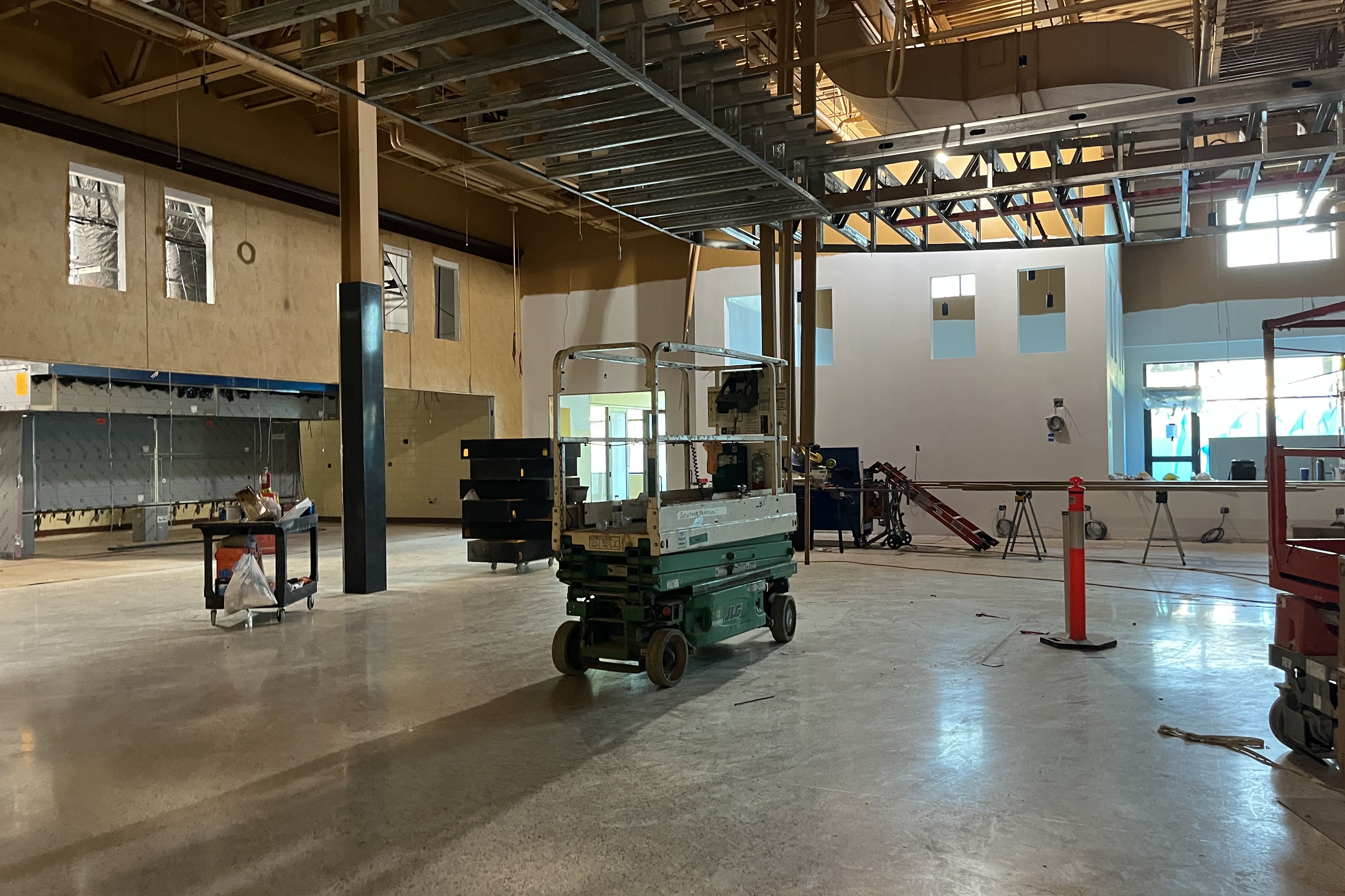 Interior construction site with exposed ceiling beams and ductwork, hanging wires; a green scissor lift, scaffold, and carts in a large room; beige walls with window openings.