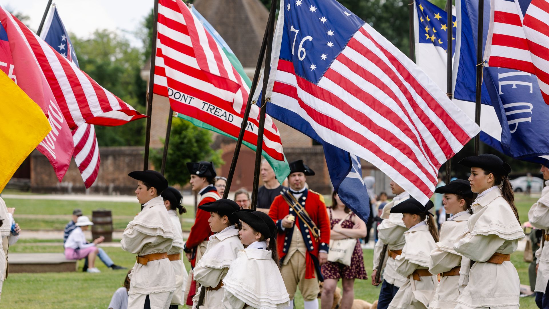 Flag-filled parade in a park with flags on poles, including the U.S. flag showing "DON'T TREAD ON ME", the Australian flag, and Union Jack. People in cream period costumes march in formation.