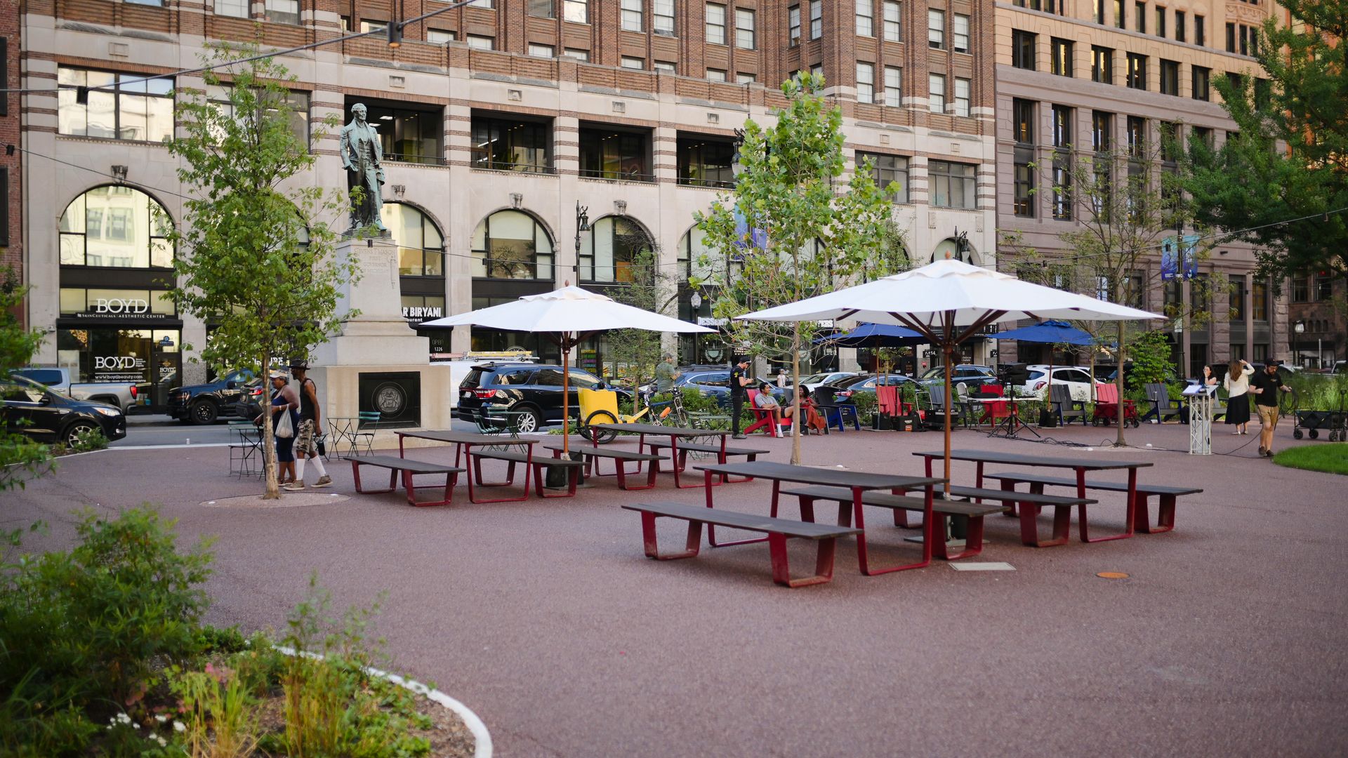 Outdoor urban space with red picnic tables under white umbrellas, trees, people walking and sitting, parked cars, and a statue in front of tall brick office buildings.