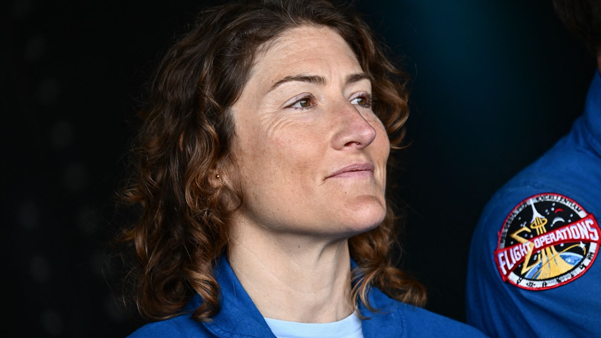 NASA Astronaut Christina Koch listens during a press conference, smiling softly in a blue flight suit.