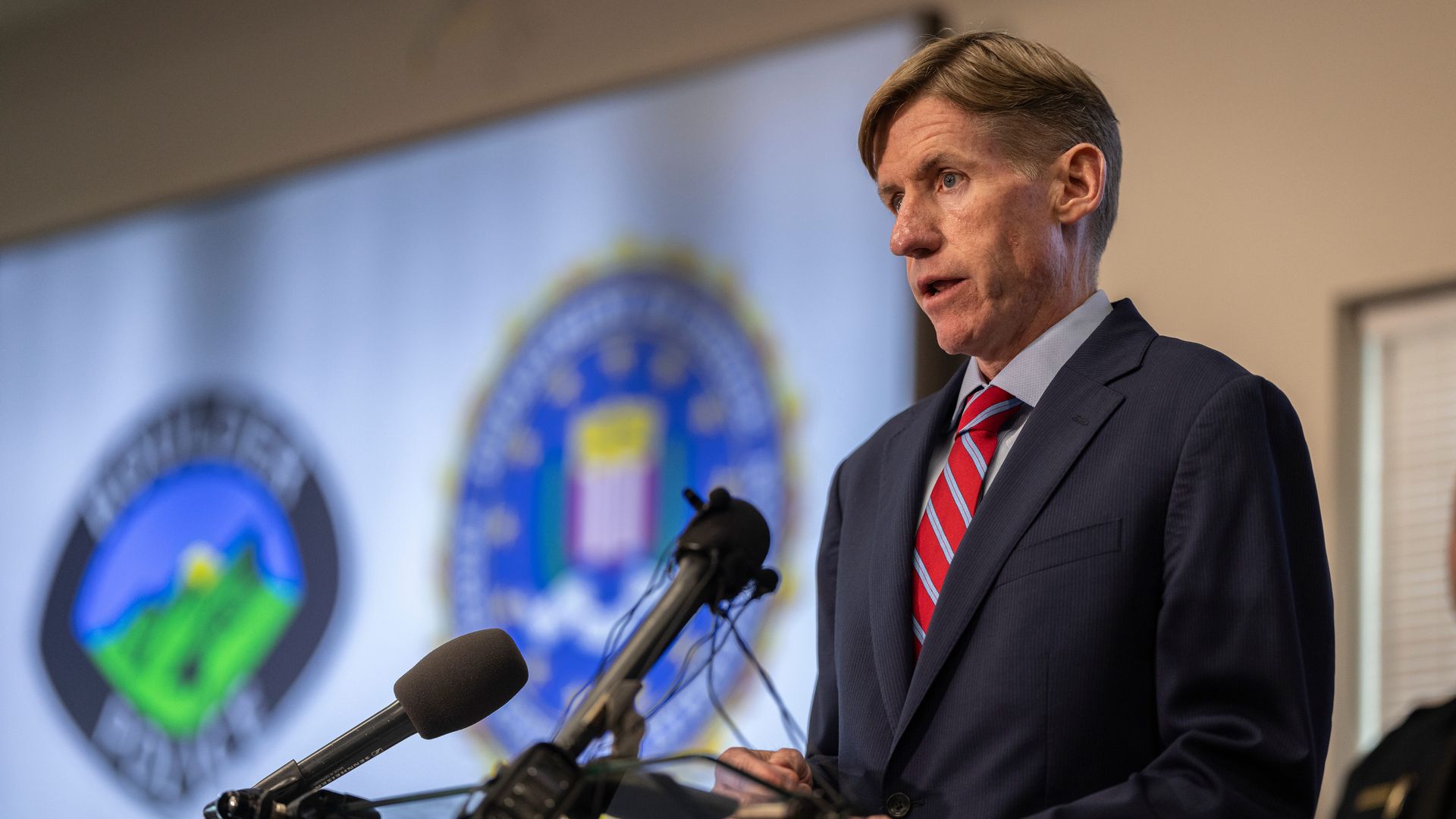 A man in a dark suit with a red striped tie speaks at a podium with several microphones during a press conference, with blurred government seals in the background.